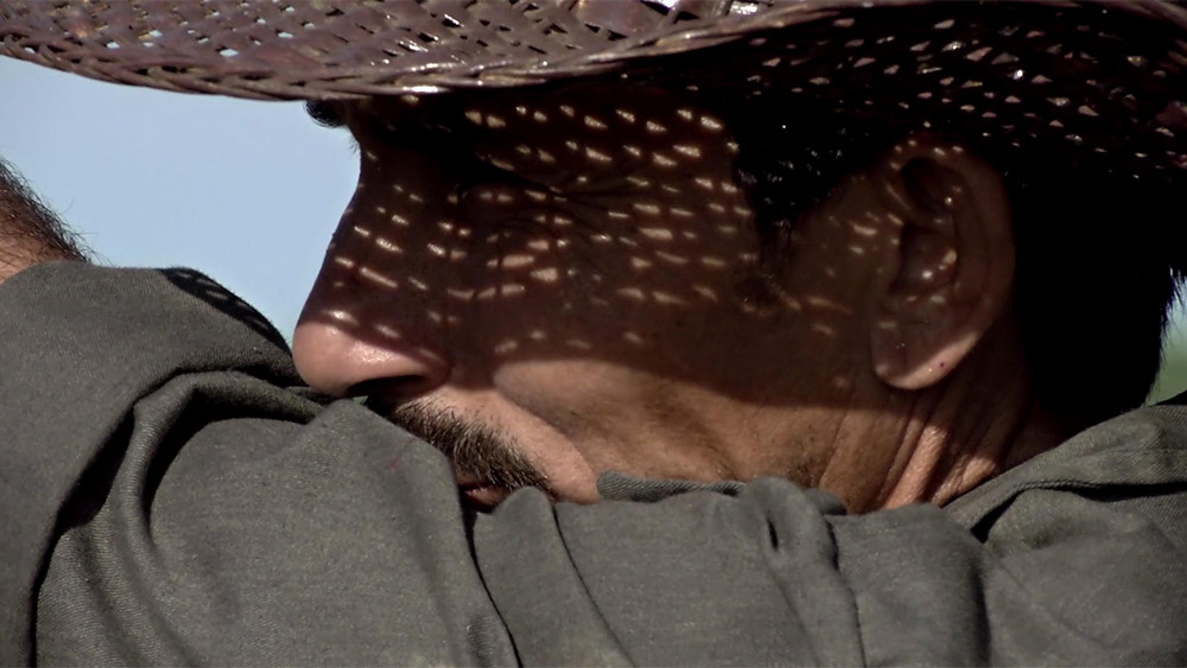 Close-up of a man’s face in partial shadow wearing a hat, his arm resting on his shoulder in the foreground.