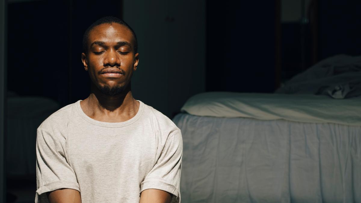 Photo of a person meditating in sunlight on the floor of a room beside a bed.