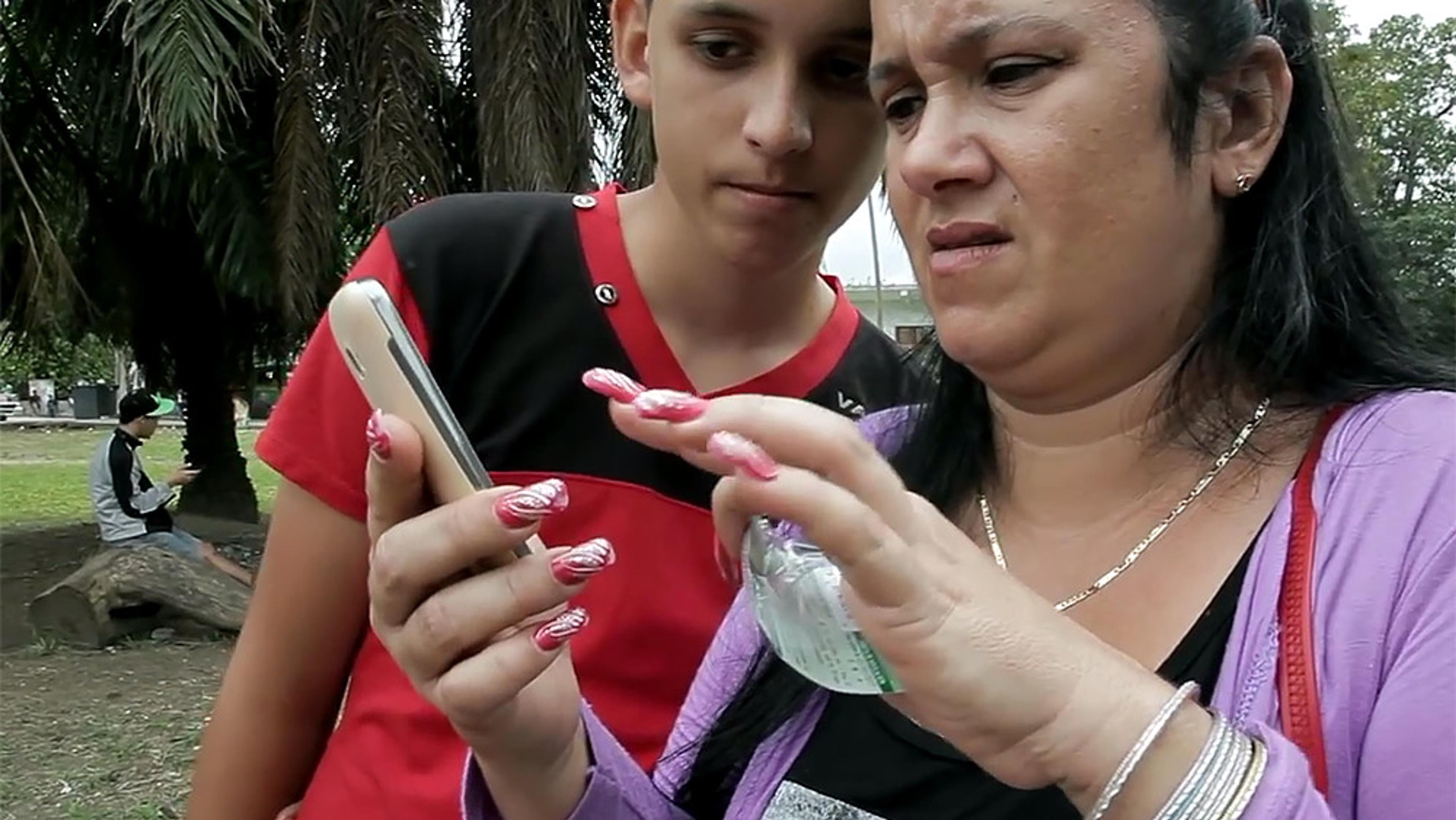 A woman and a boy outdoors, both looking at a smartphone. The woman’s expression is confused.