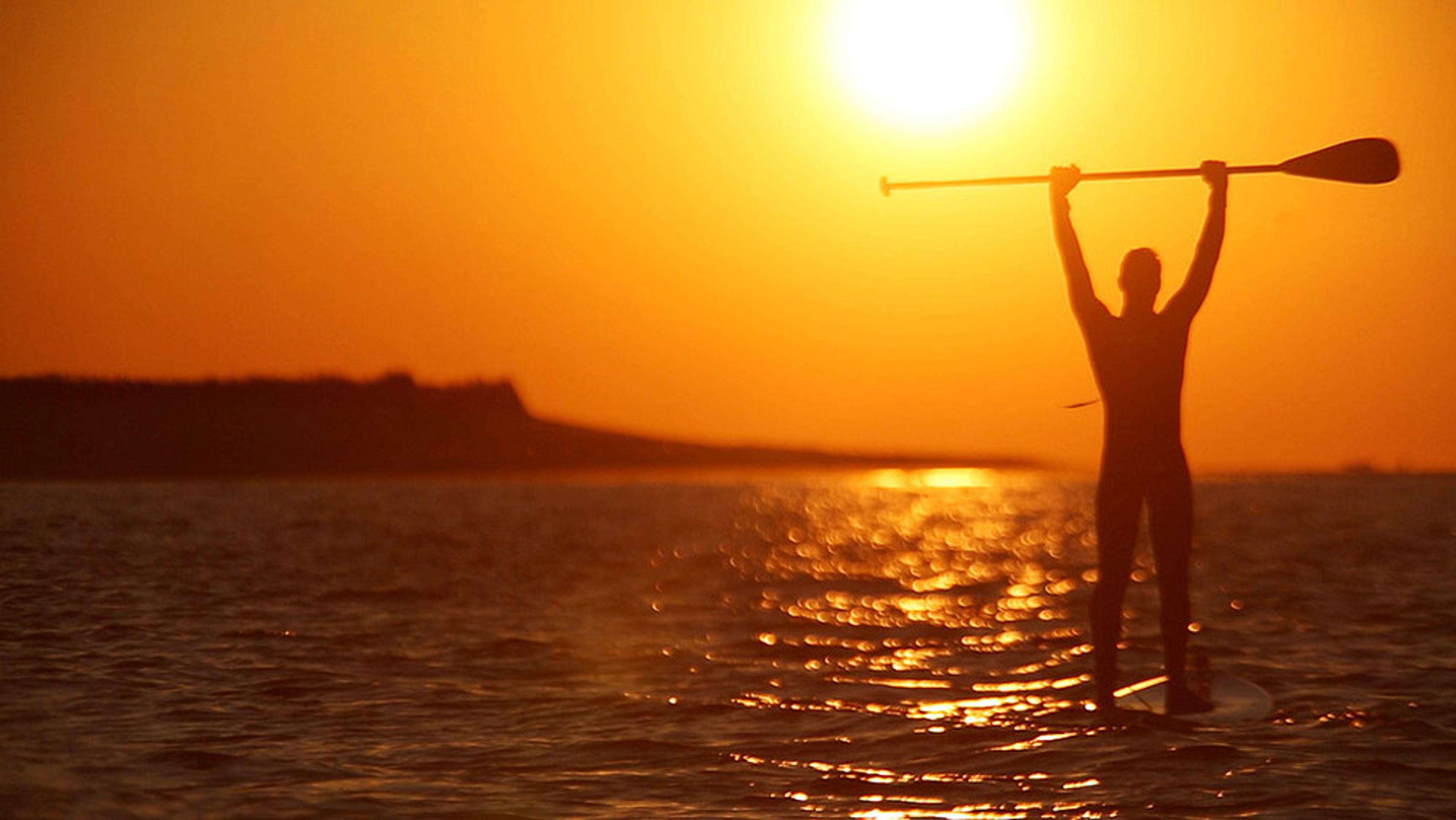 Photo of a man on a paddleboard holding a paddle overhead, silhouetted against a golden sunset over calm water.
