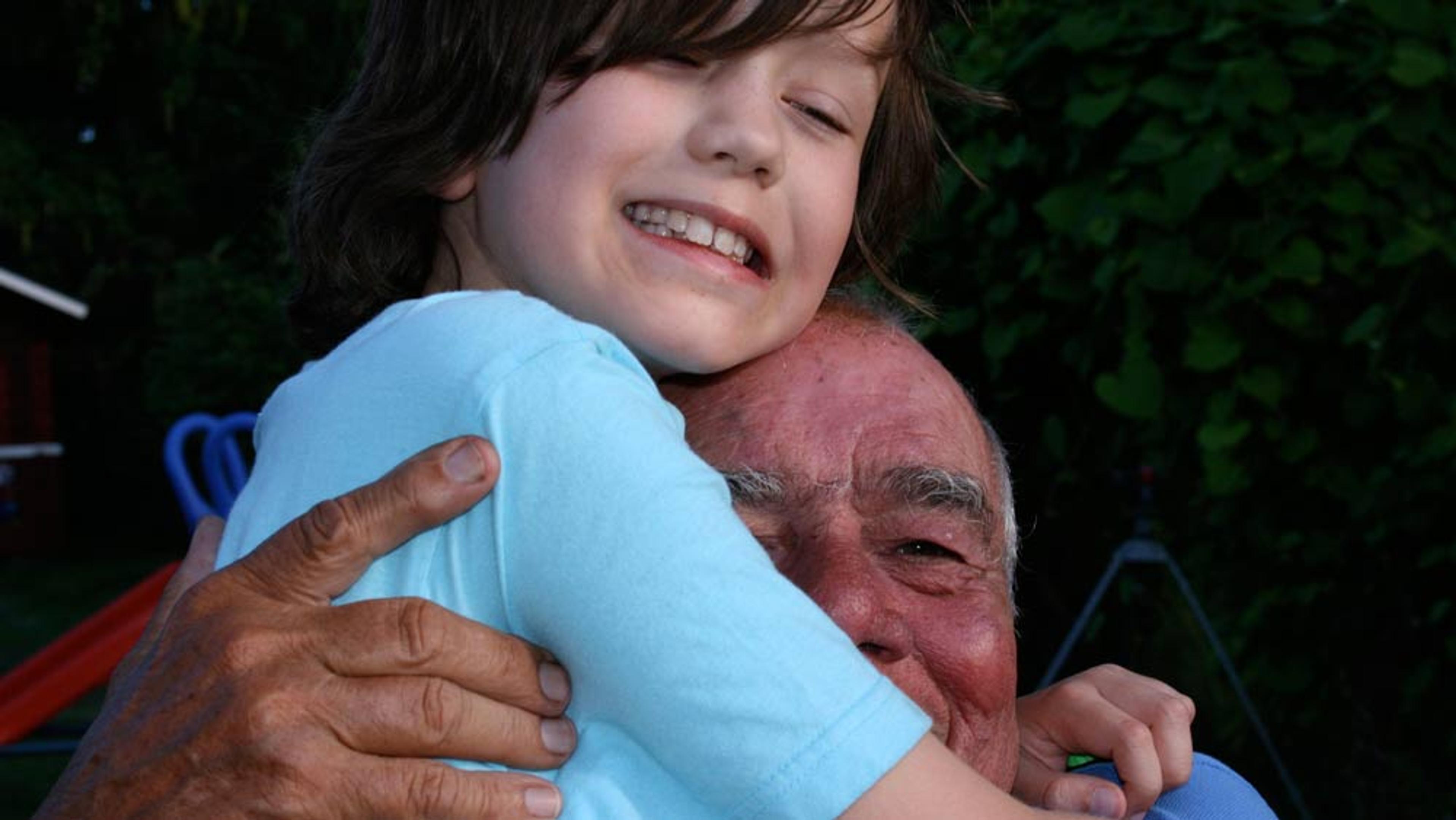 A child hugging an elderly man outdoors, both smiling with eyes closed, surrounded by greenery in the background.