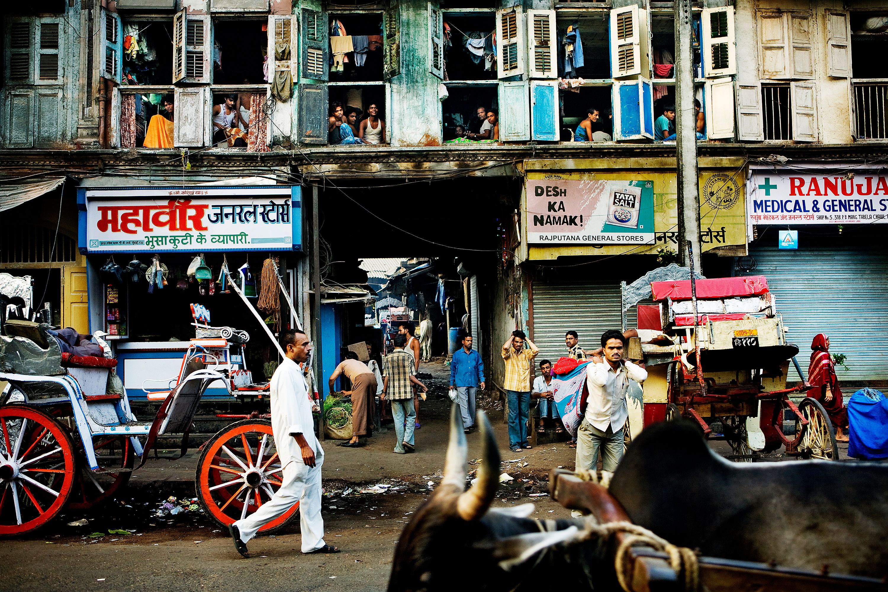 Street scene in India with people, bullock carts and shops, showing a bustling atmosphere.