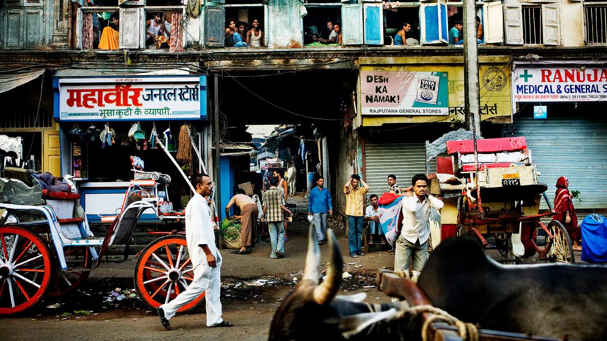 Street scene in India with people, bullock carts and shops, showing a bustling atmosphere.