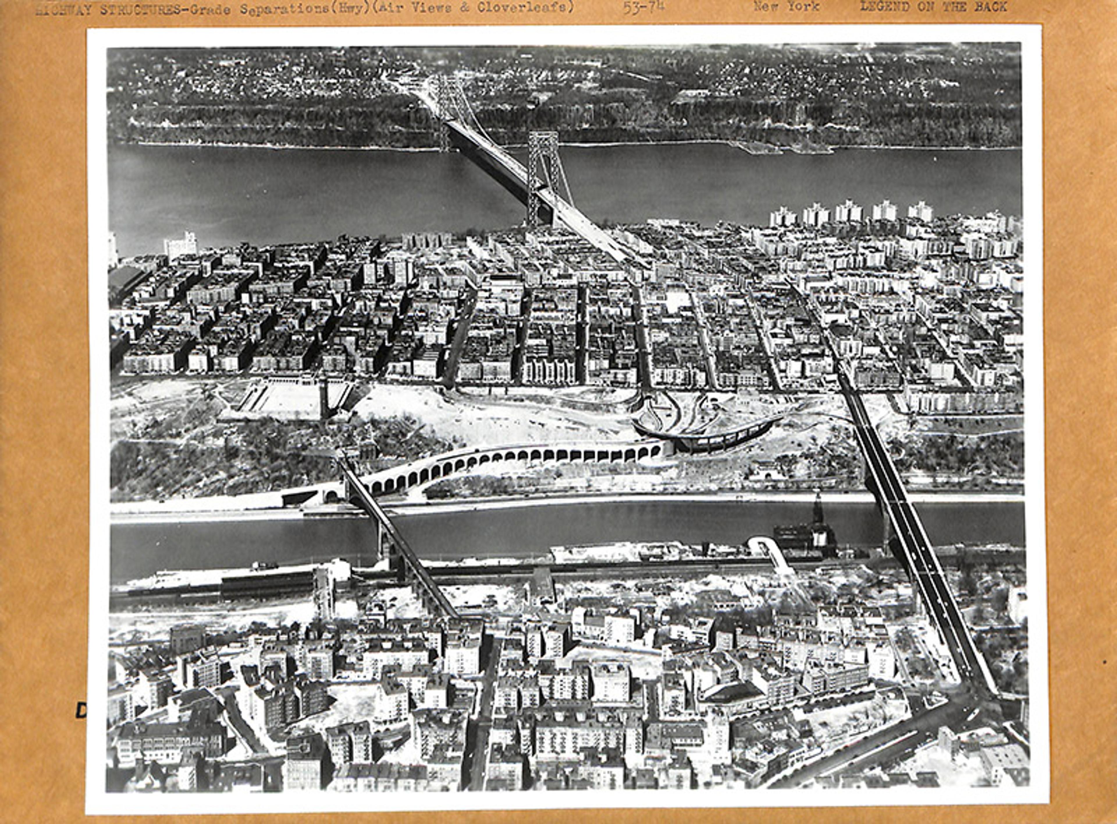 A black and white aerial photo of a cityscape with a large suspension bridge crossing a river and urban buildings below.