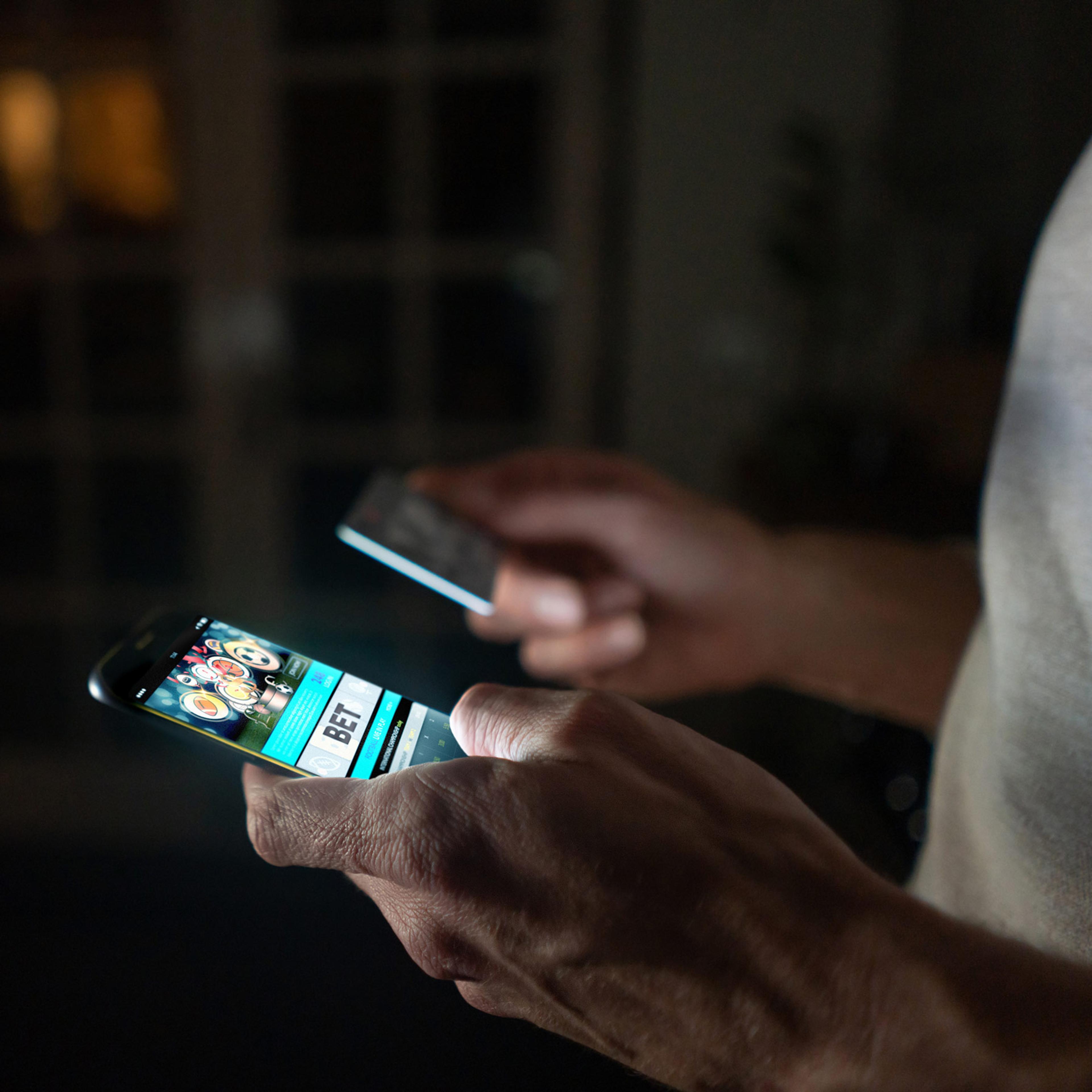A close-up of a man using an online betting platform on his smartphone. The phone screen lights up a darkened room