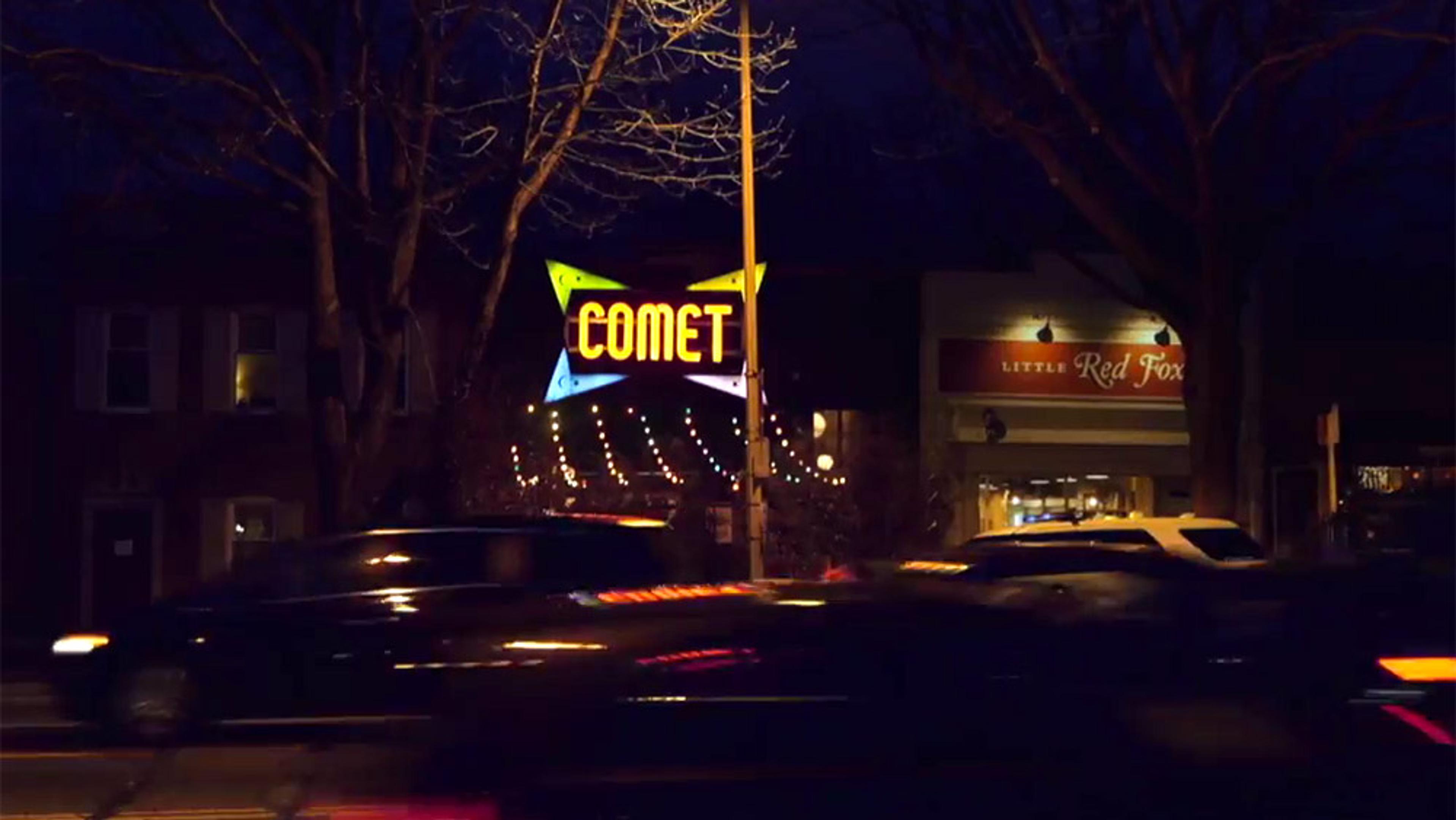 A neon sign reading “COMET” on a building facade at night, with cars passing in front and a “Little Red Fox” shop nearby.