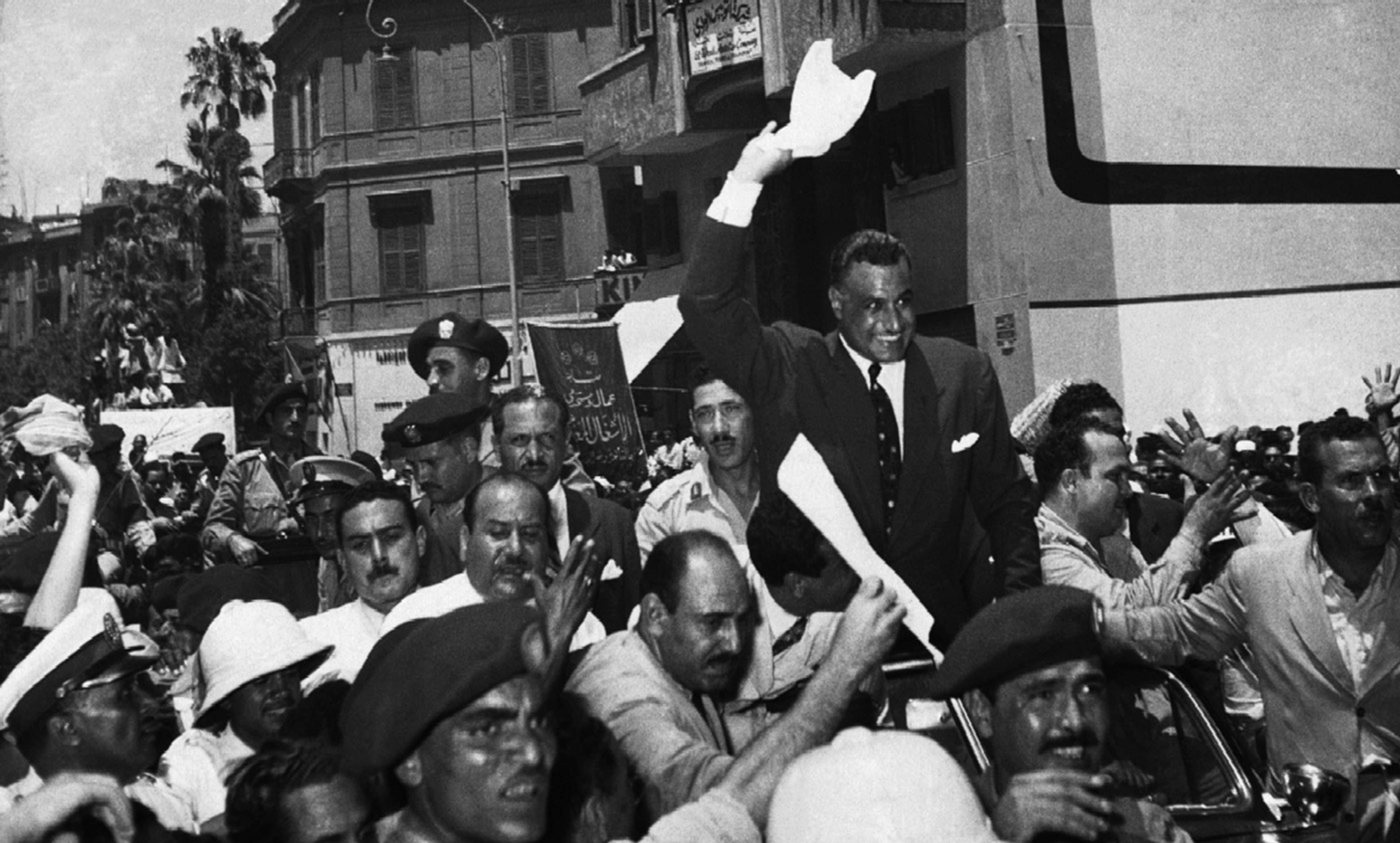 Black and white photo of a man in a suit, surrounded by a large crowd and waving, in an urban setting with buildings in the background.