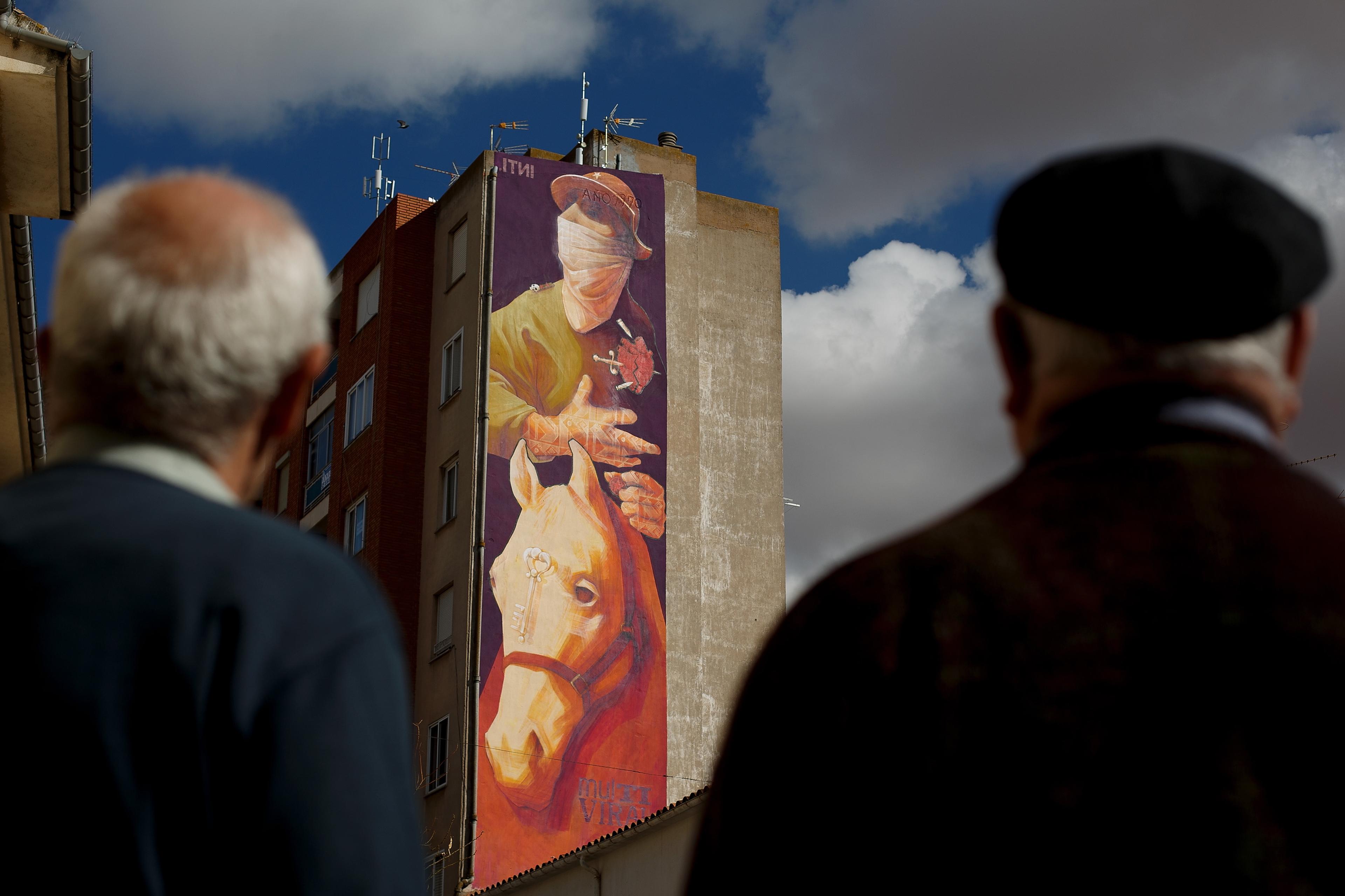 Photo of a mural depicting a person and a horse on a tall building with two men in the foreground looking up.