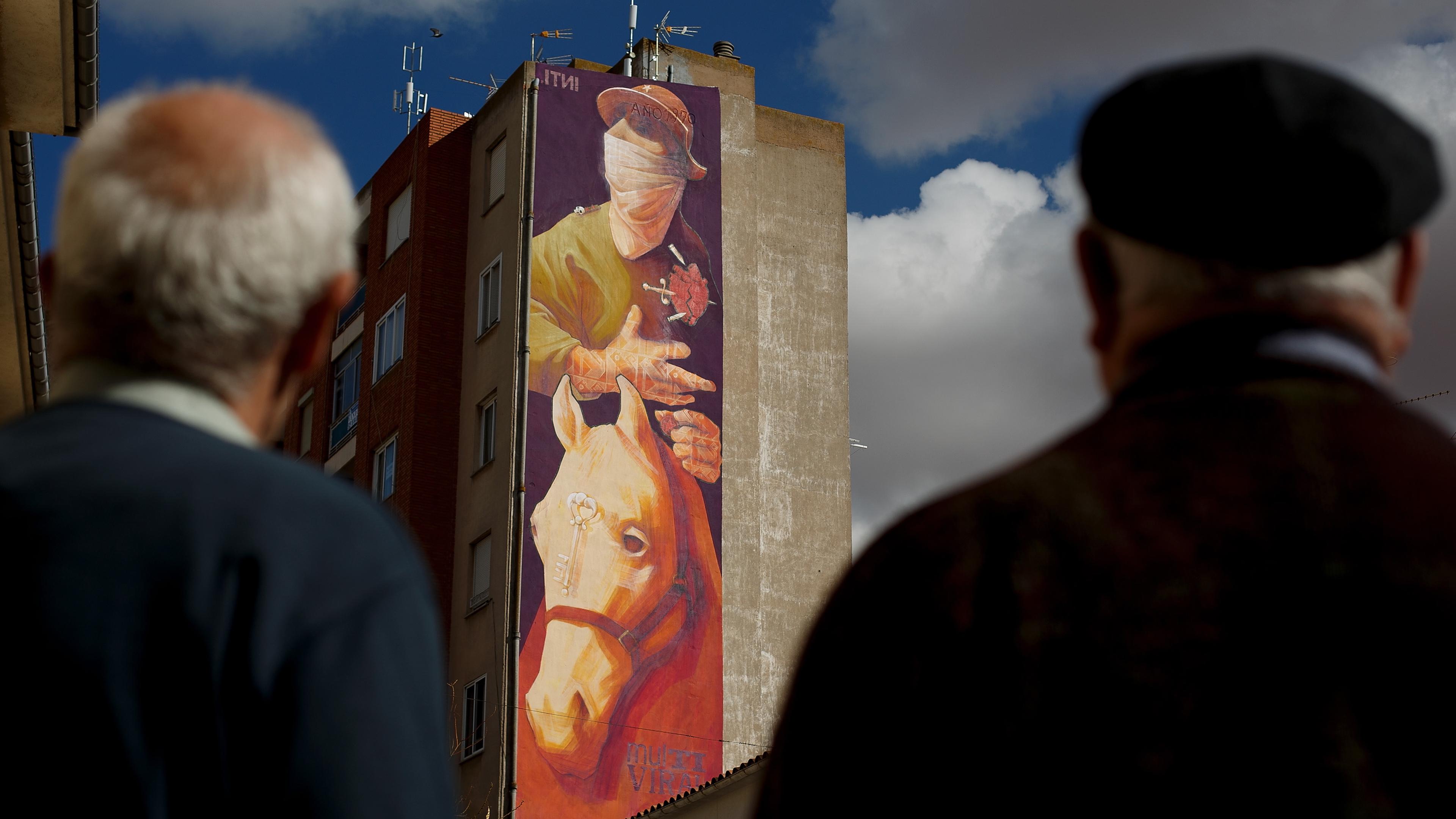 Photo of a mural depicting a person and a horse on a tall building with two men in the foreground looking up.