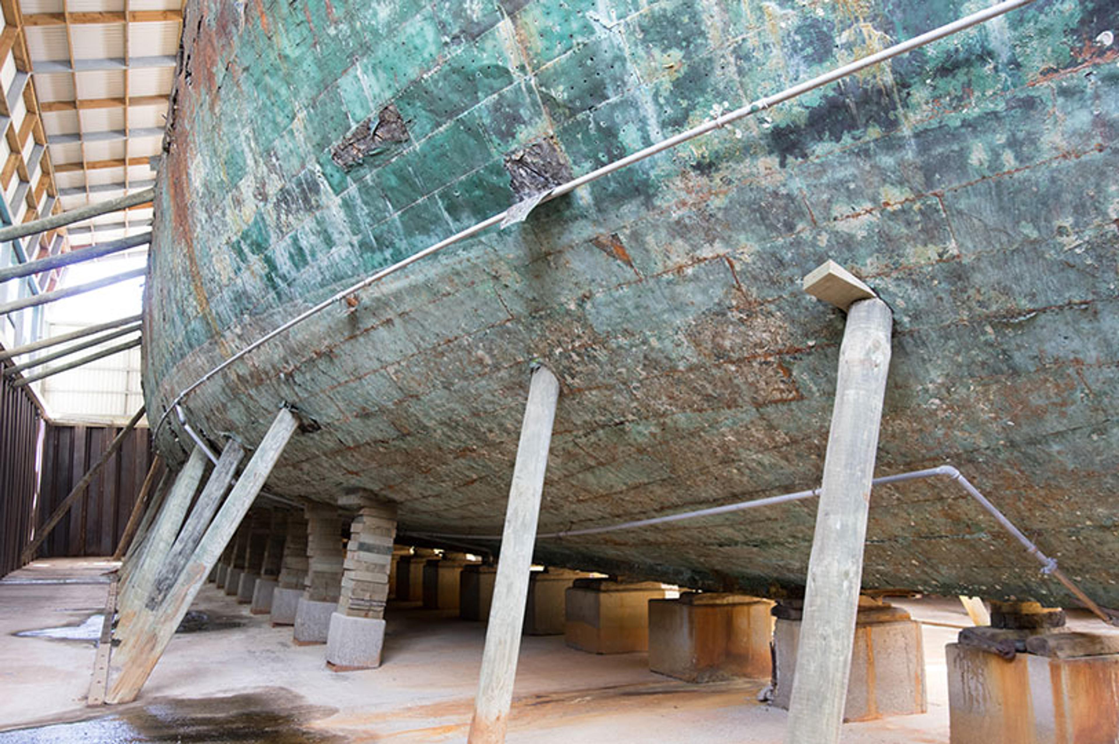The bottom of an old, weathered ship’s hull supported by wooden and concrete stands in a covered drydock.