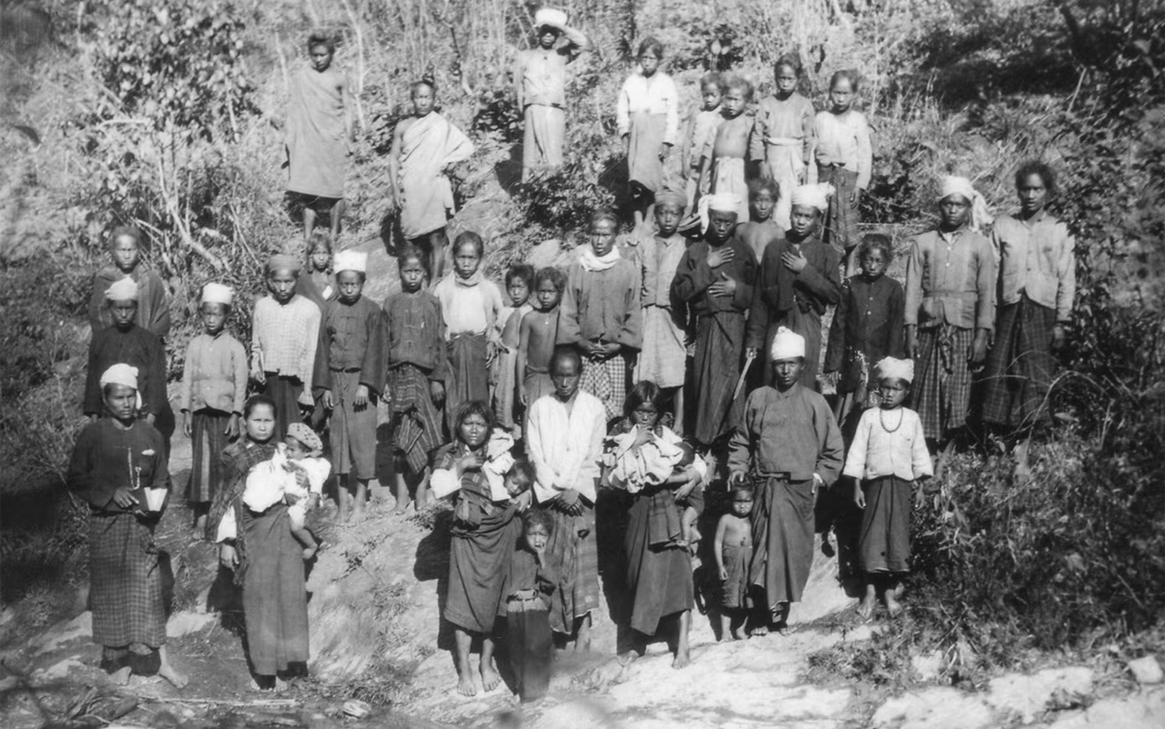 Black and white photo of a group of people standing outdoors on a slope by a pond with trees and bushes in the background.