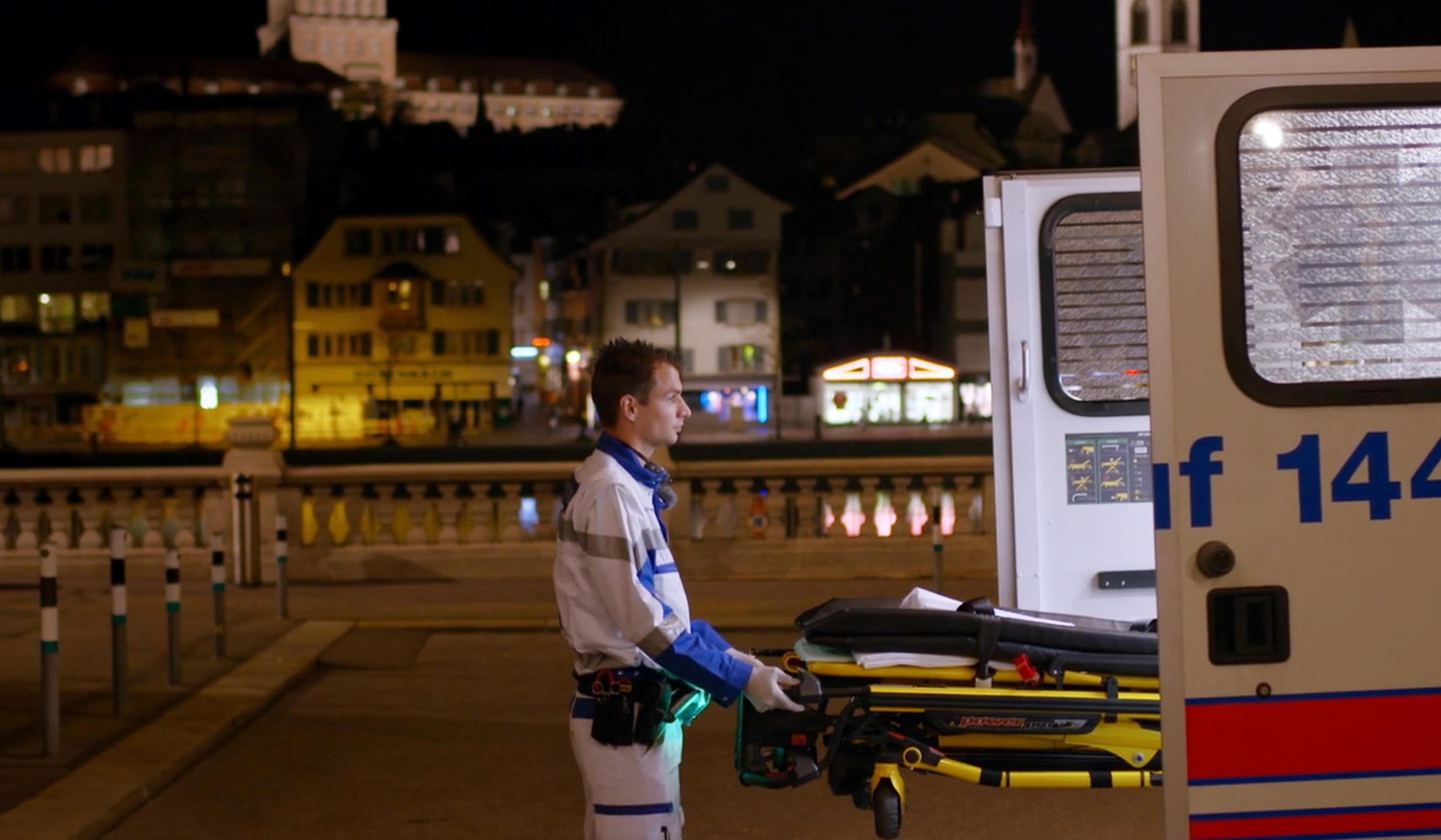 Paramedic loading a stretcher into an ambulance at night with lighted buildings in the background.