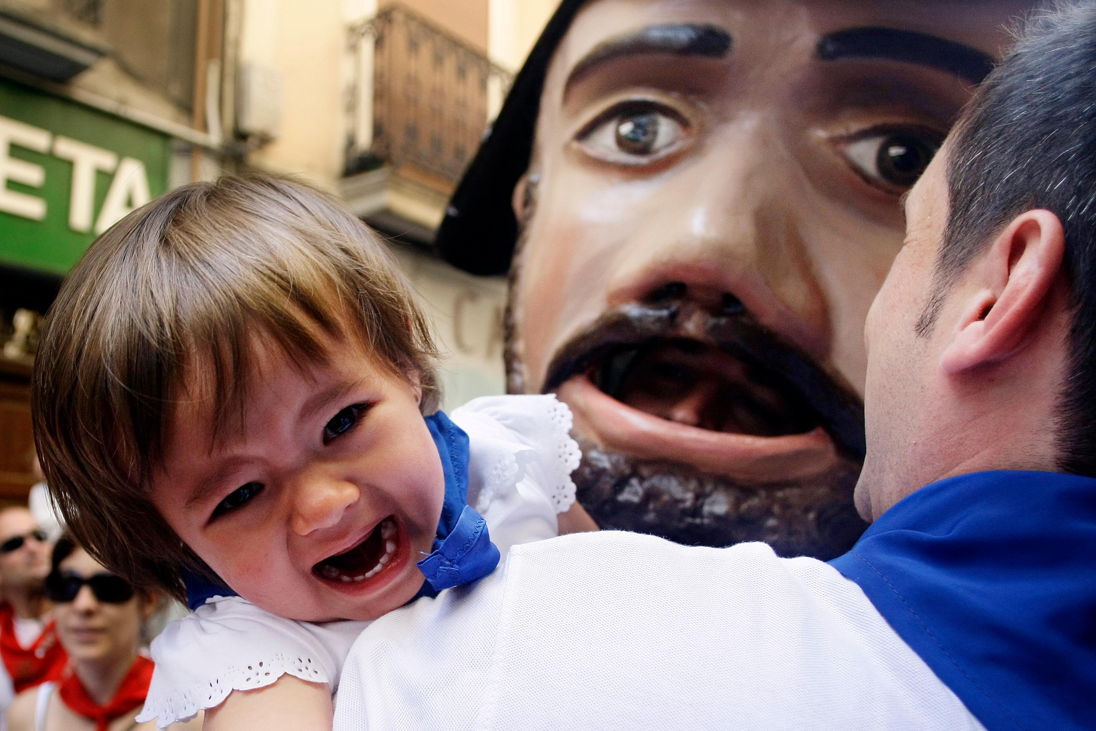 Photo of a crying child with an adult near a giant mask in a street setting. The child wears a blue scarf and white clothes.