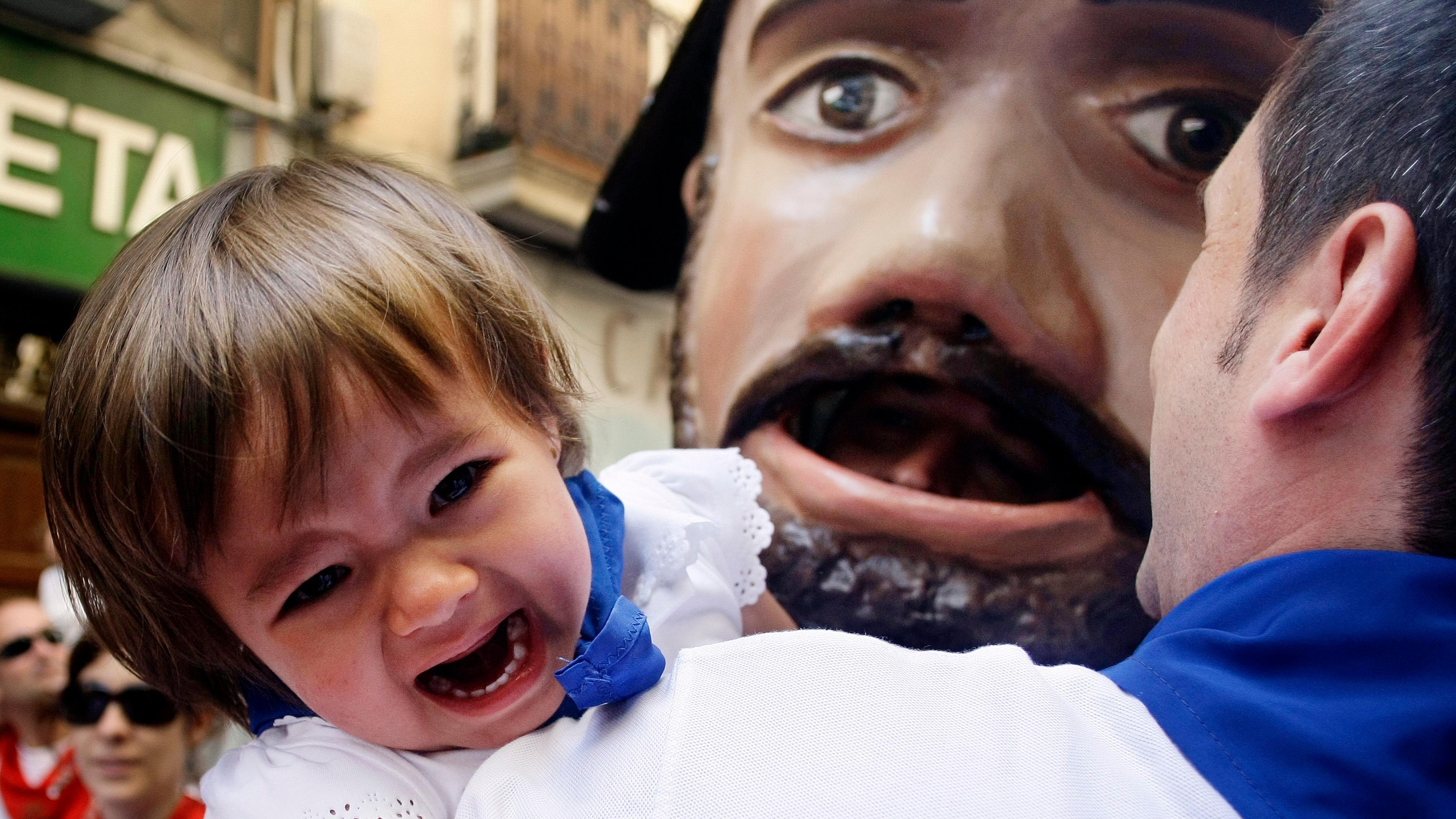 Photo of a crying child with an adult near a giant mask in a street setting. The child wears a blue scarf and white clothes.