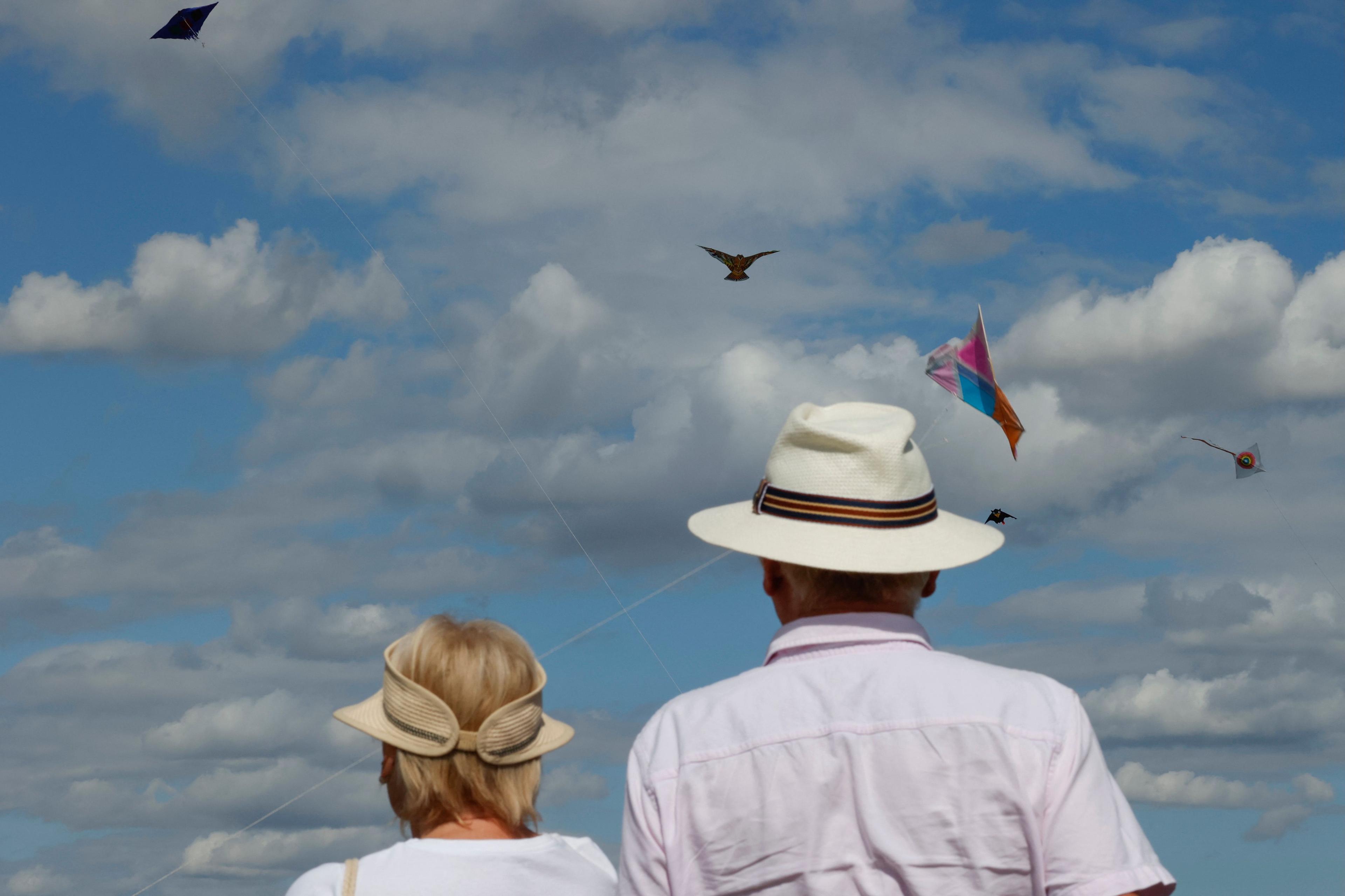 Photo of two people in hats watching colourful kites in a blue sky with fluffy clouds.