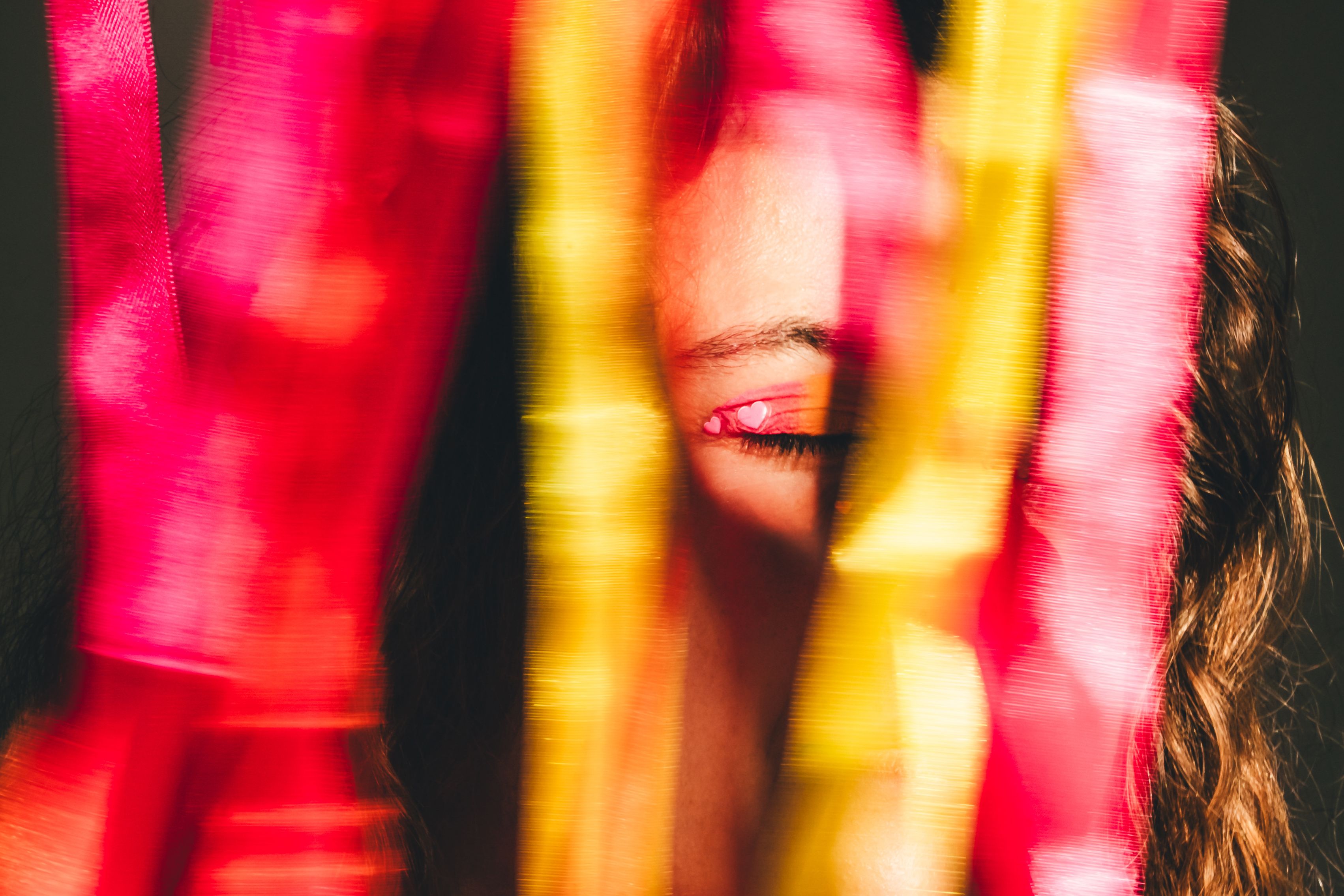 Photo of a person’s closed eyes with colourful ribbons in red and yellow partially obscuring the face.