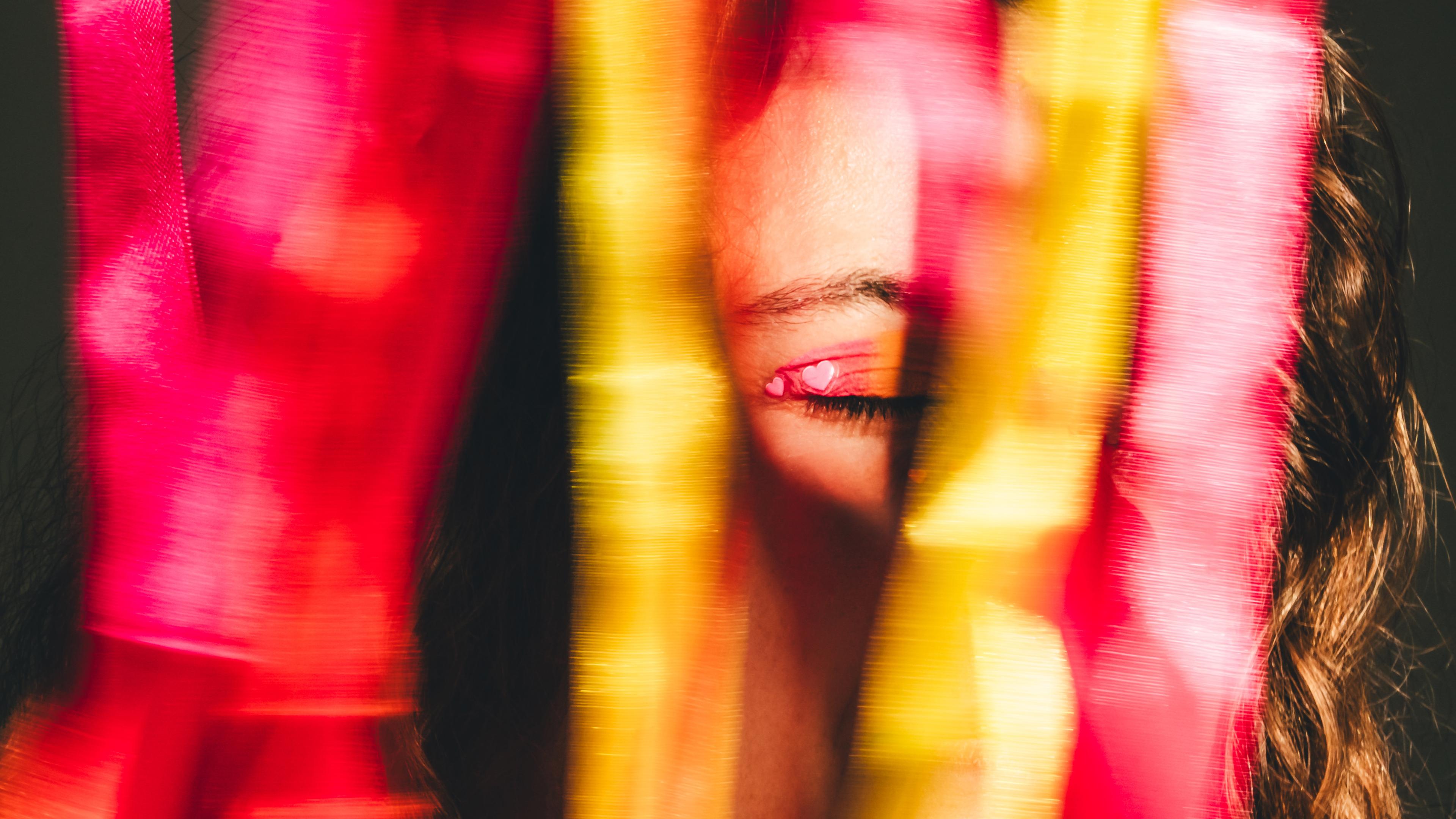 Photo of a person’s closed eyes with colourful ribbons in red and yellow partially obscuring the face.