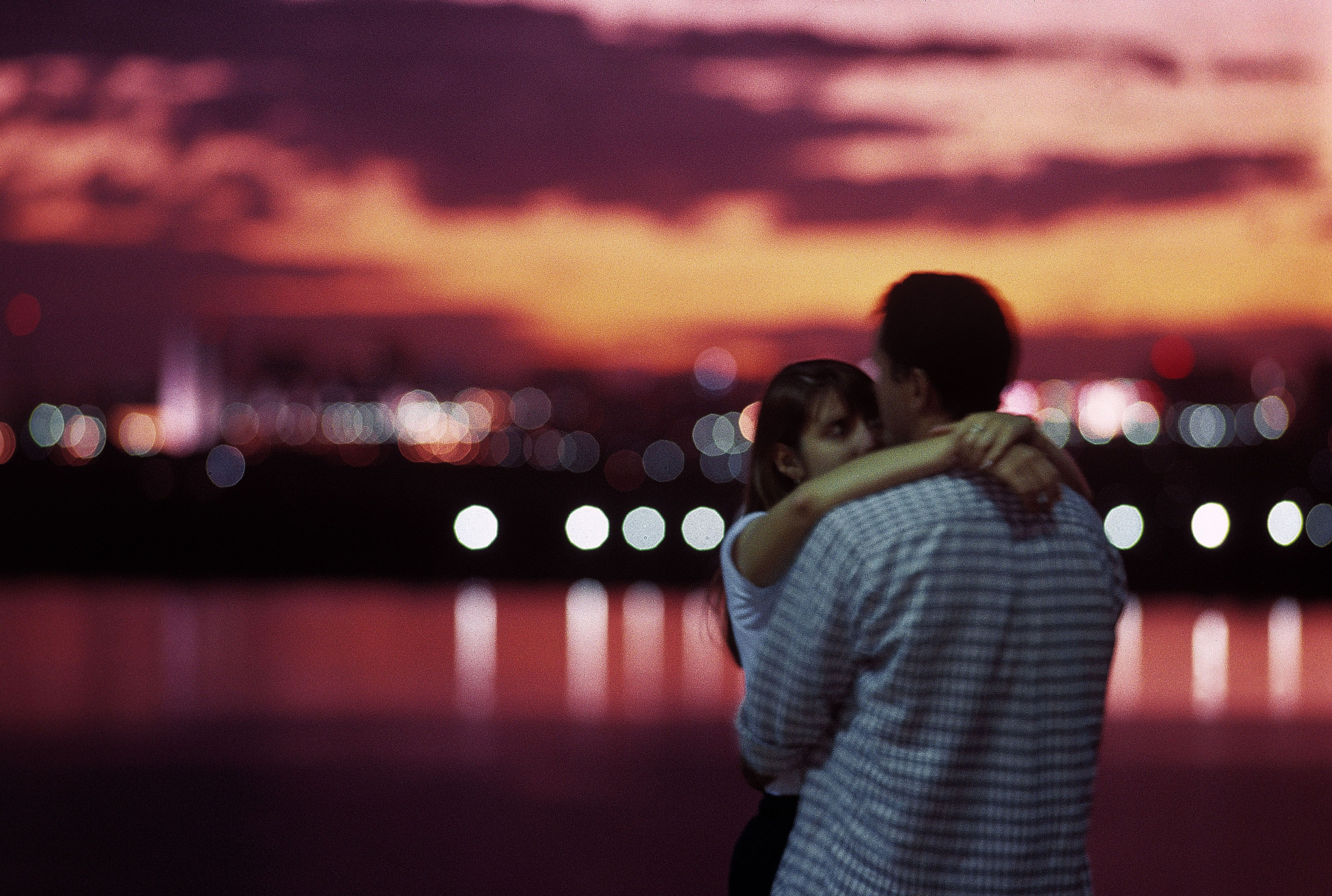Photo of a couple embracing at sunset with a blurred cityscape and colourful sky in the background.