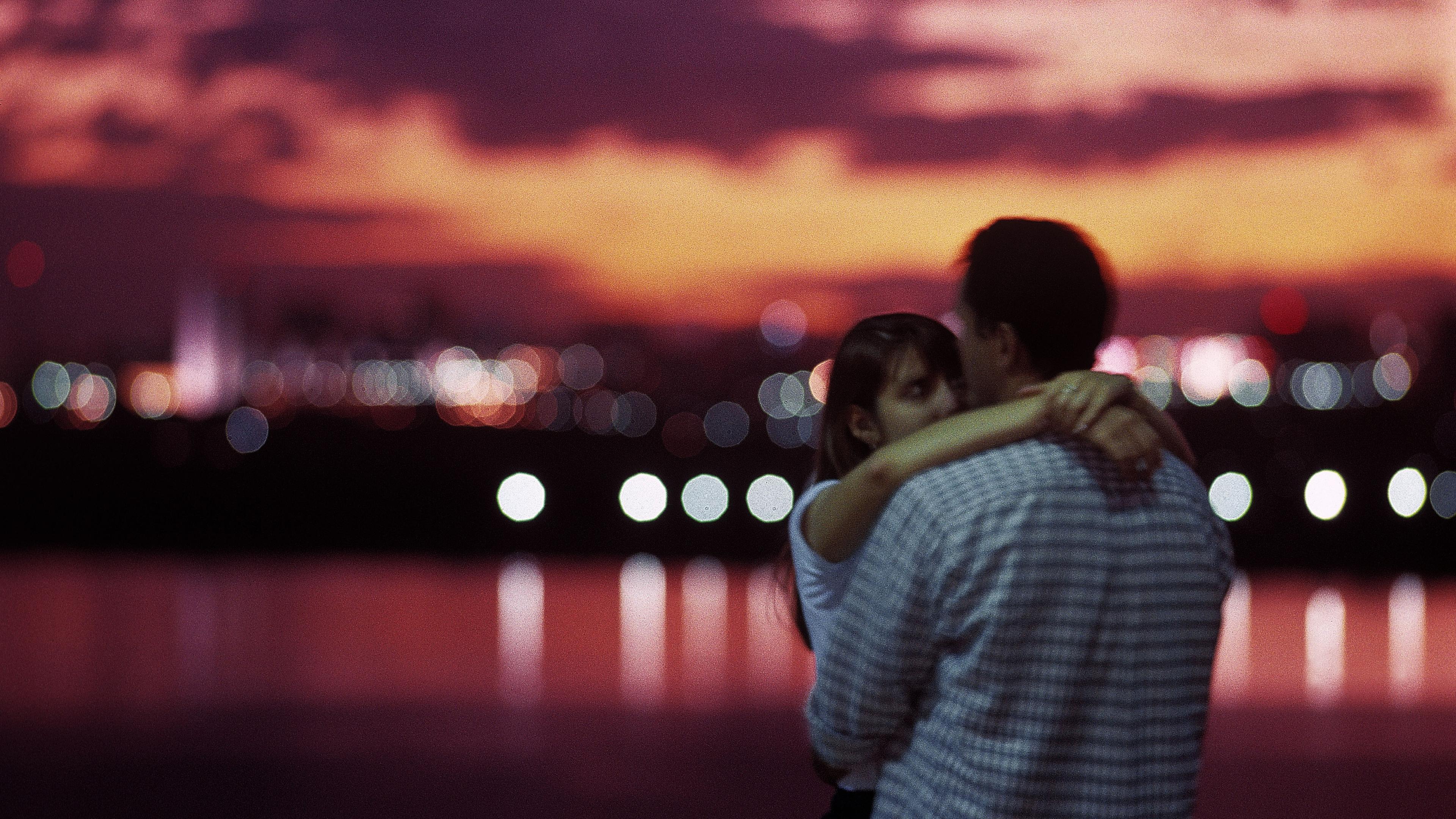 Photo of a couple embracing at sunset with a blurred cityscape and colourful sky in the background.