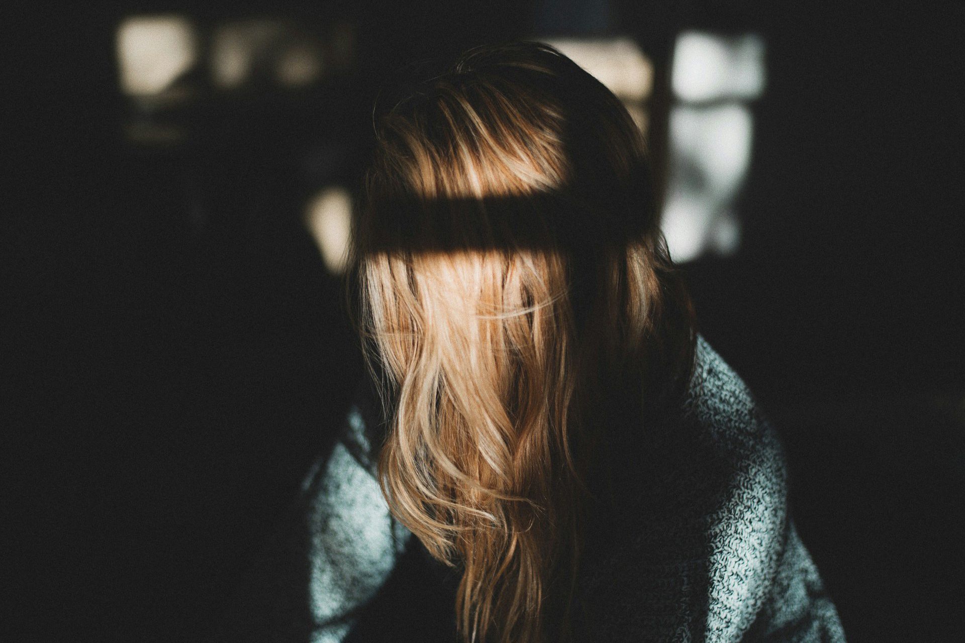 A person with long blonde hair covering they’re face in shadowy light.