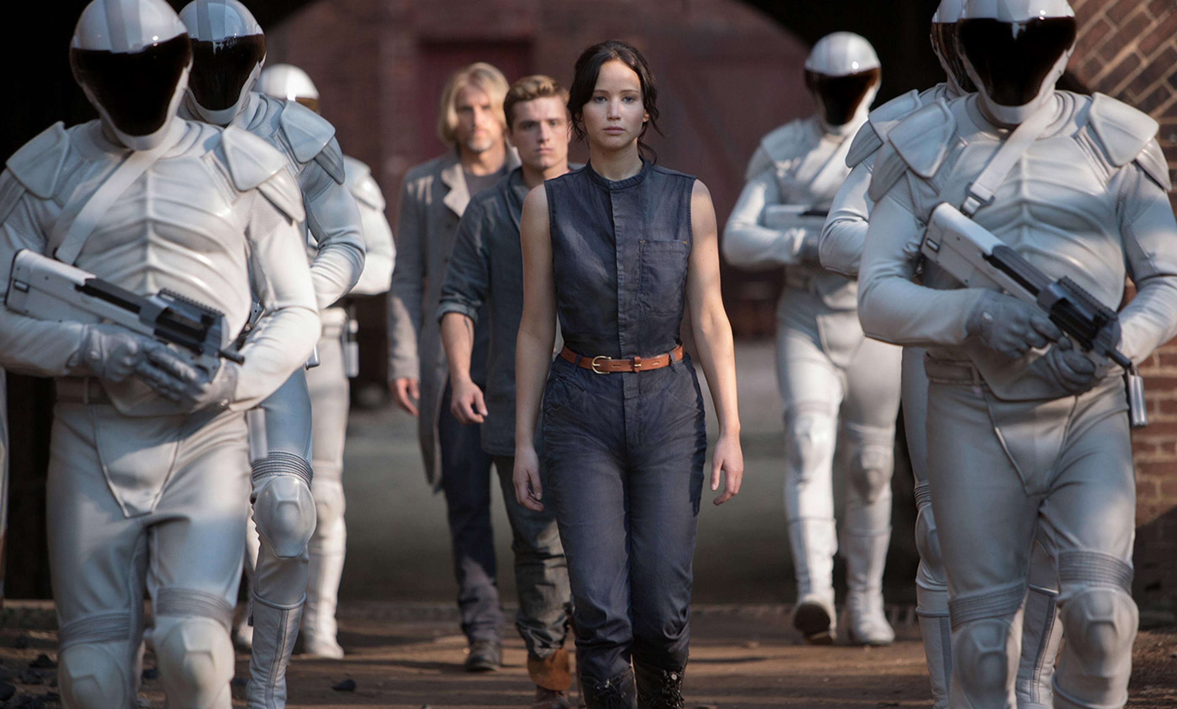 Film still of a woman in a jumpsuit who is Jennifer Lawrence as Katniss walking flanked by armoured guards with helmets and white suits.