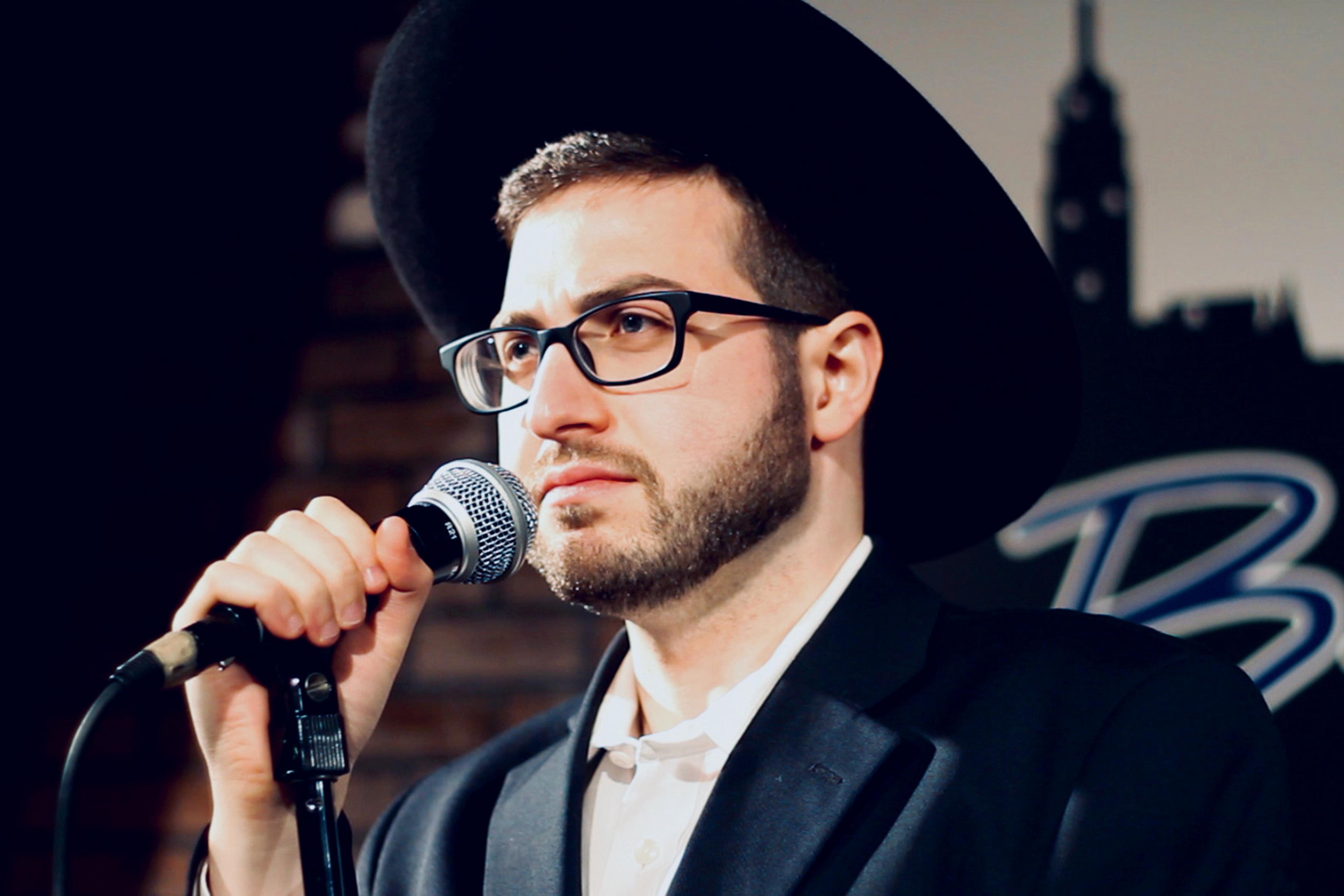 A Jewish man wearing glasses and a platchige biber hat, speaking into a microphone at a comedy club, with a brick wall and a blurred sign in the background.