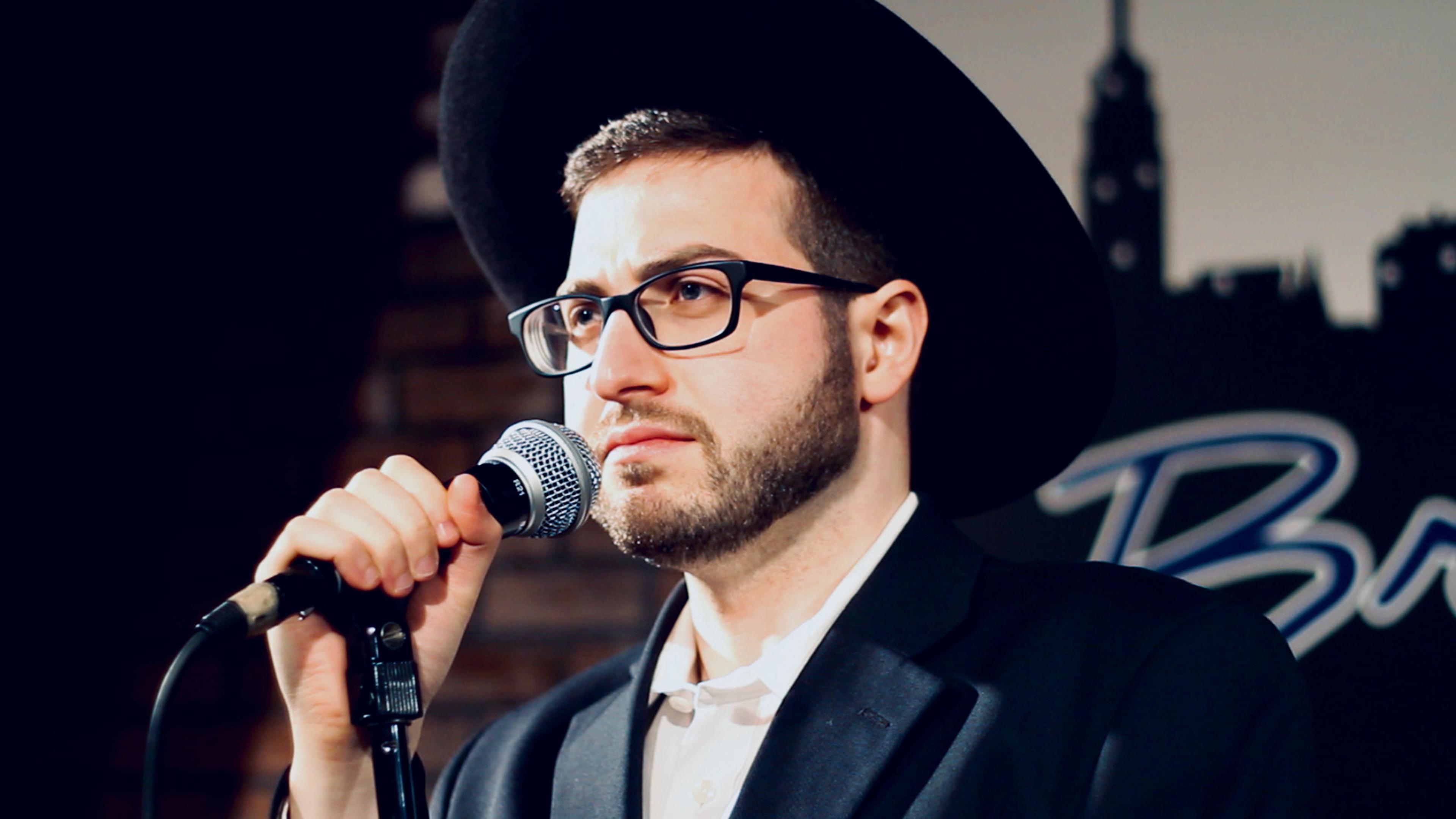 A Jewish man wearing glasses and a platchige biber hat, speaking into a microphone at a comedy club, with a brick wall and a blurred sign in the background.