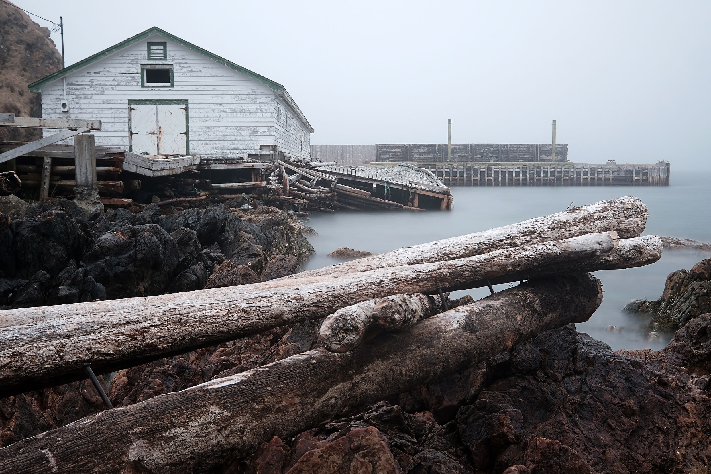 An old wooden building and jetty on a rocky shoreline with logs in the foreground and misty water in the background.