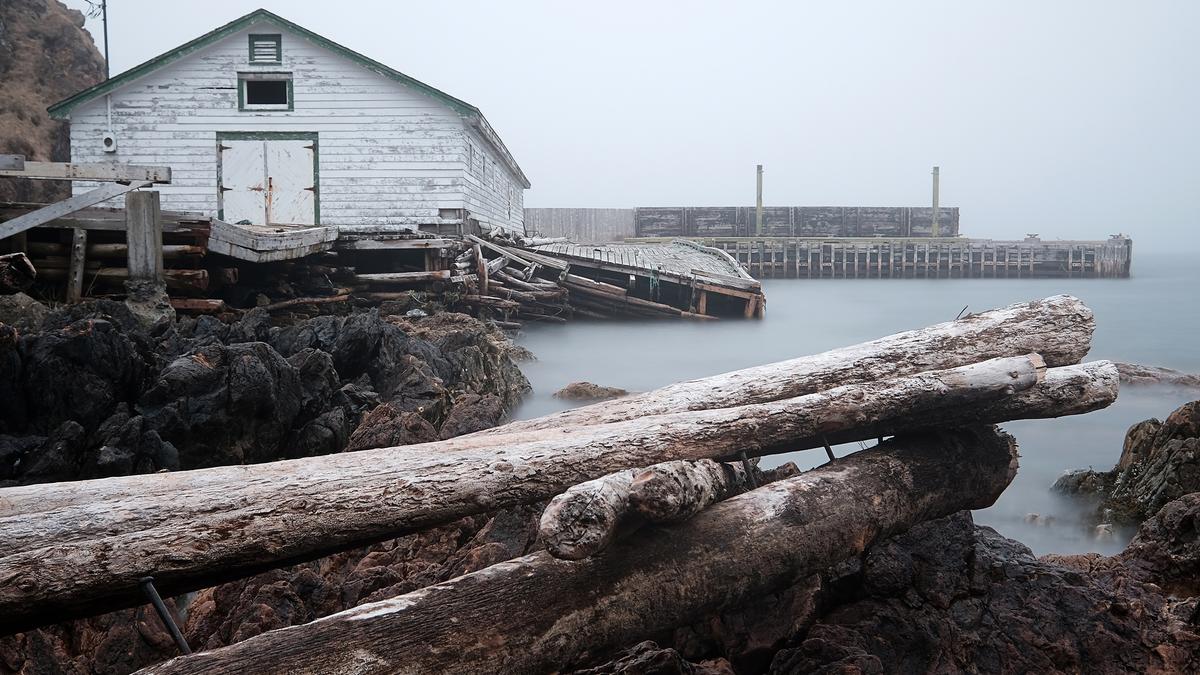 An old wooden building and jetty on a rocky shoreline with logs in the foreground and misty water in the background.