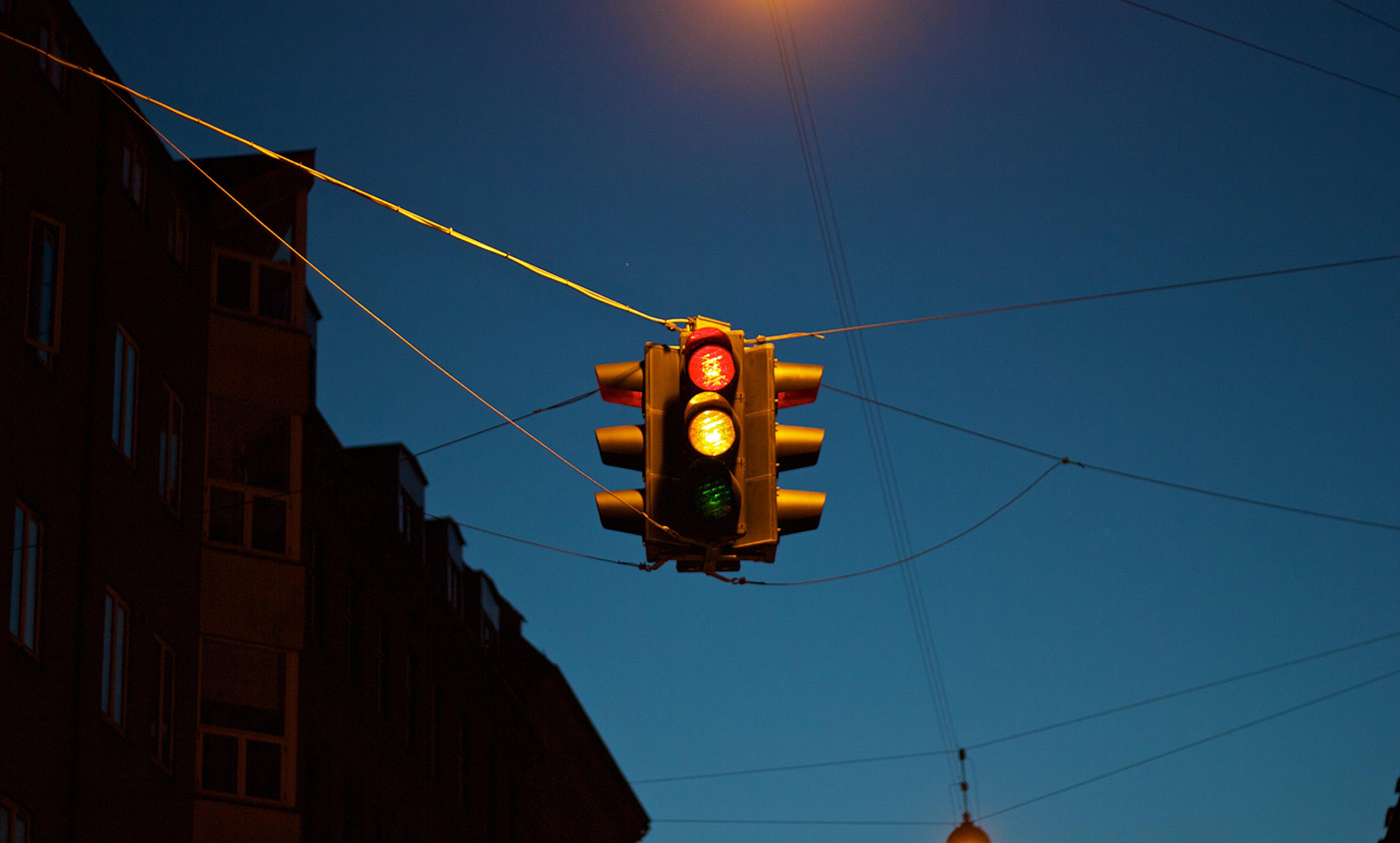 A traffic light hanging in front of a dark building at dusk, showing red, yellow and green lights with visible cables.