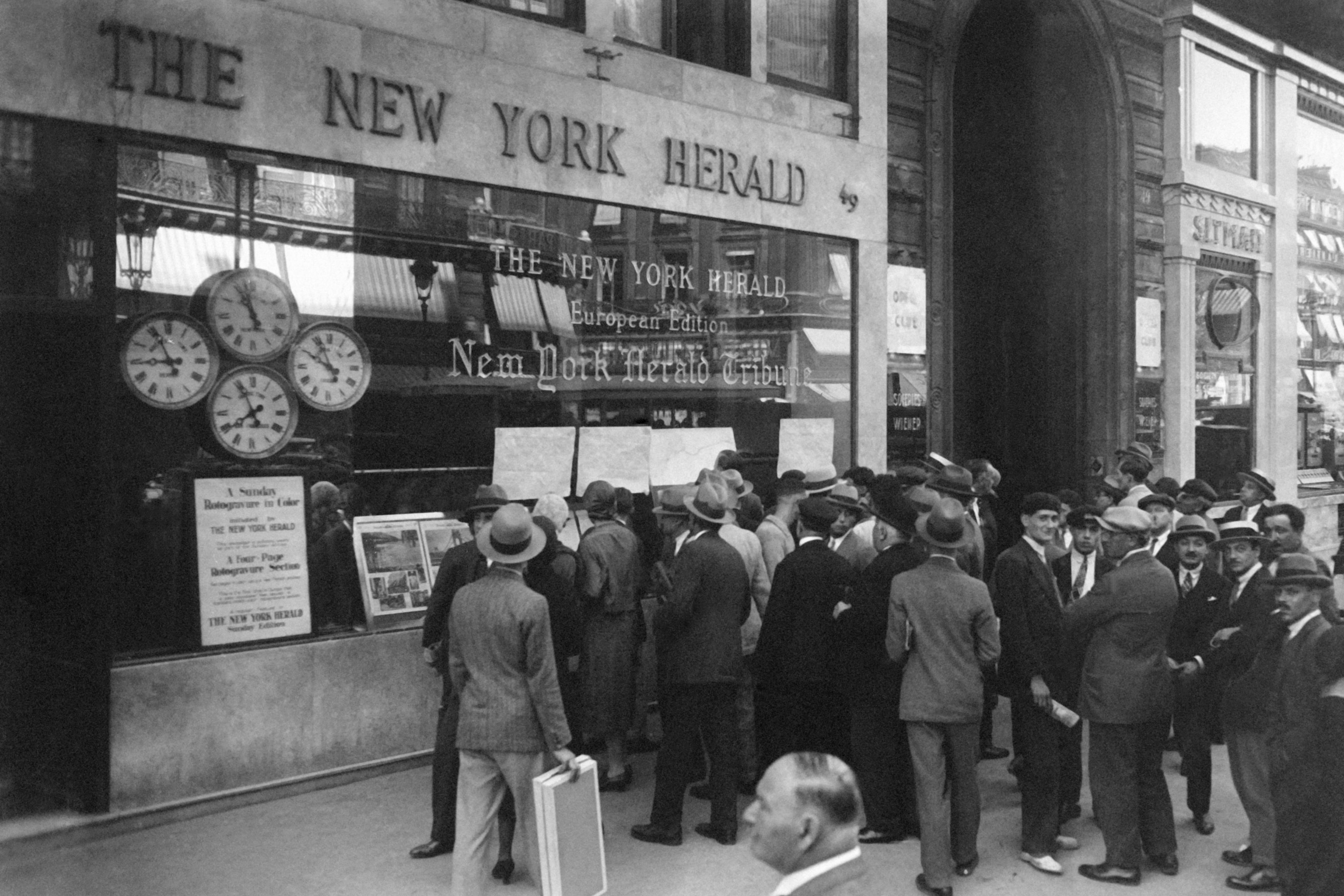 Black and white photo of a crowd outside The New York Herald building reading posted newspapers and looking at a clock display.