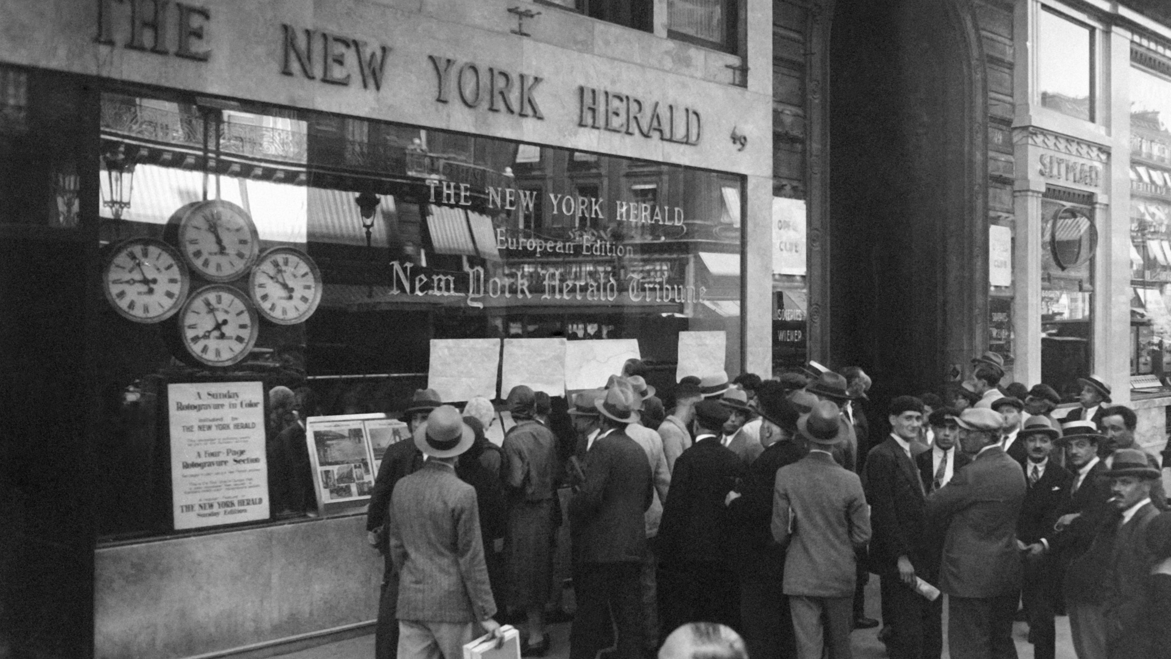 Black and white photo of a crowd outside The New York Herald building reading posted newspapers and looking at a clock display.