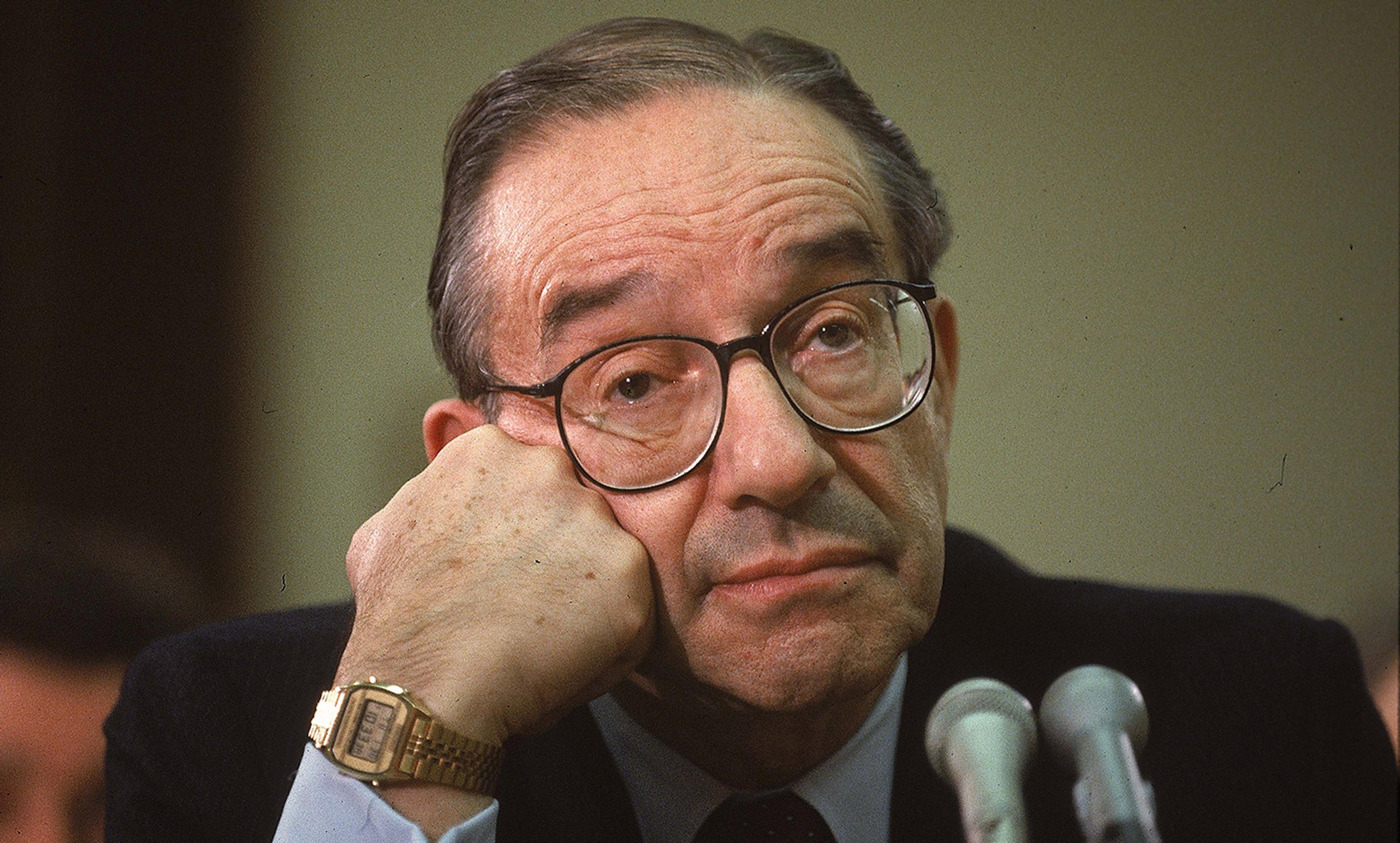 A man wearing glasses and a suit, resting his chin on his hand, looking pensive, with a gold watch visible on his wrist.
