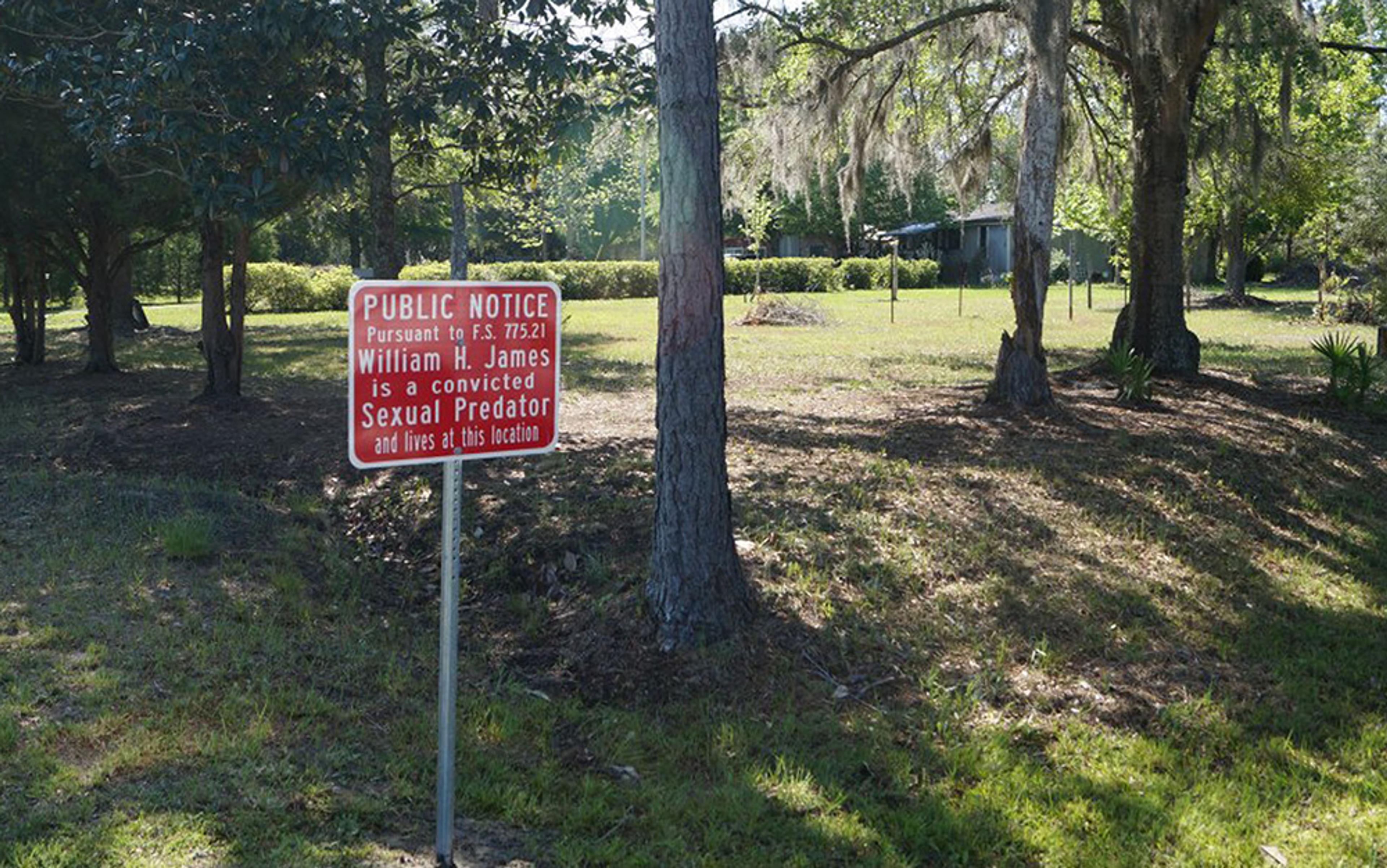 A public notice sign in a grassy area with trees, stating someone is a convicted sexual predator living at that location.