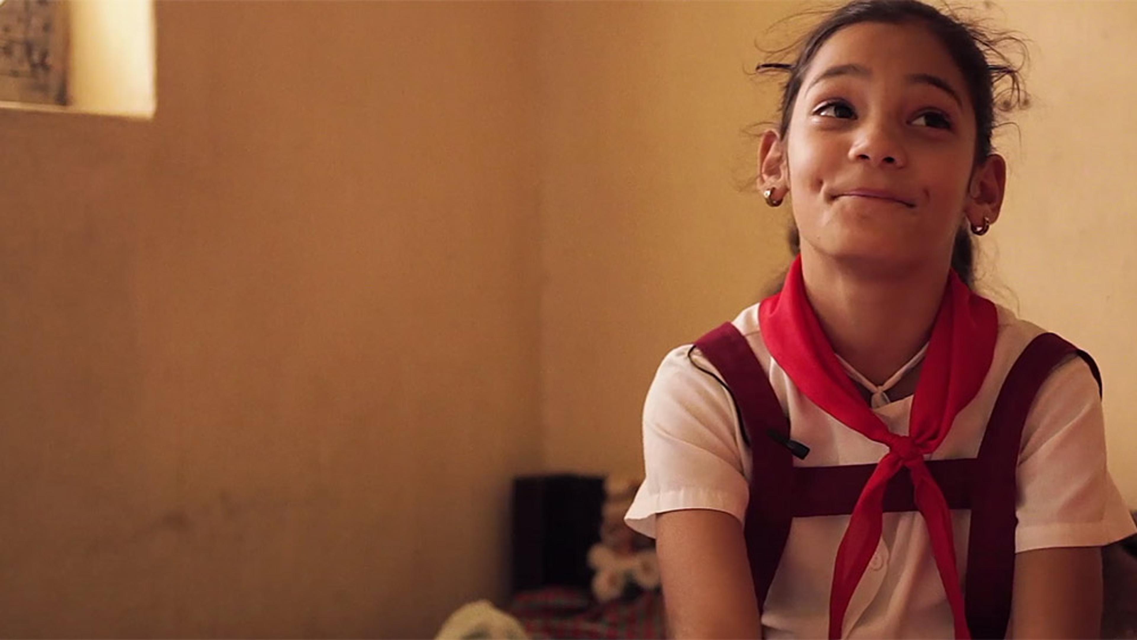 A young girl in a school uniform with a red scarf, smiling and looking upwards in a warm, softly lit room.