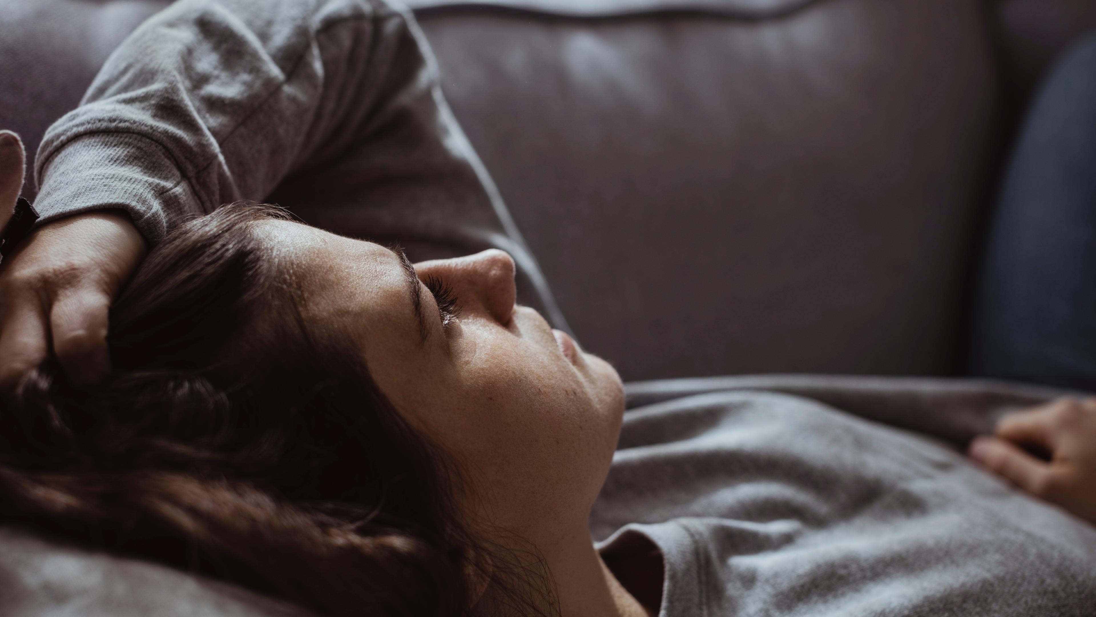 Photo of a person lying on a sofa, looking relaxed with eyes closed. They are wearing a grey jumper and holding their hair.