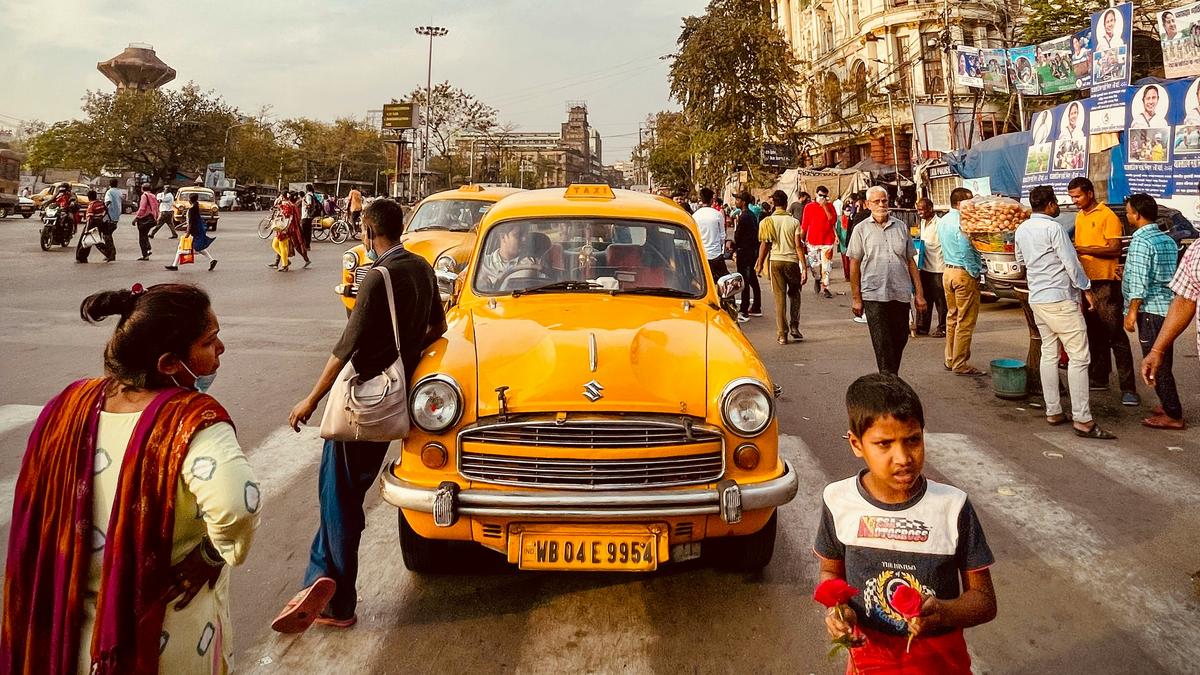 A yellow taxi on a busy street, people walking, child holding flowers, historic building in the background under a blue sky.