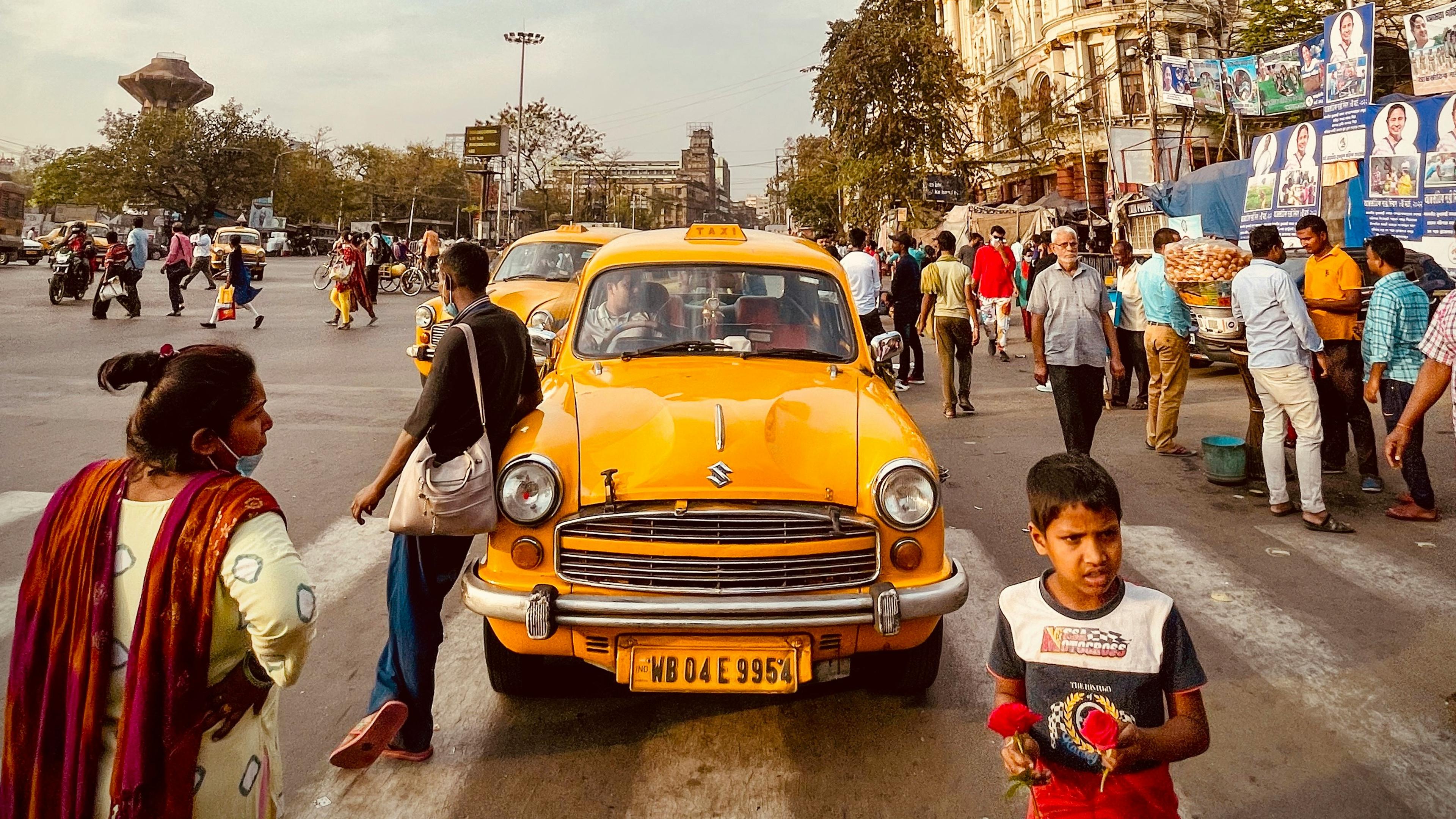 A yellow taxi on a busy street, people walking, child holding flowers, historic building in the background under a blue sky.