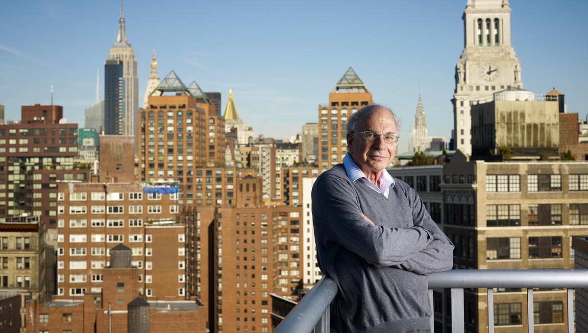 Daniel Kahneman standing on a balcony, arms crossed, with New York City skyline and buildings in the background under a clear blue sky.
