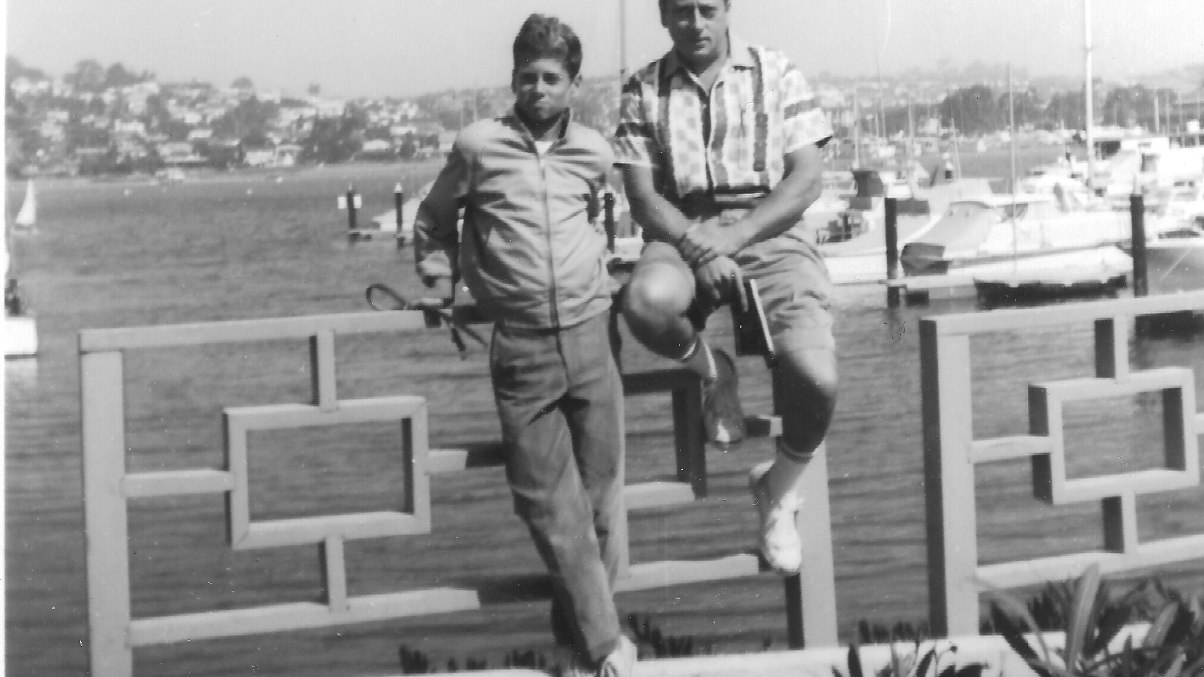 Black and white photo of two men by a waterfront fence, with boats and a hillside town in the background.