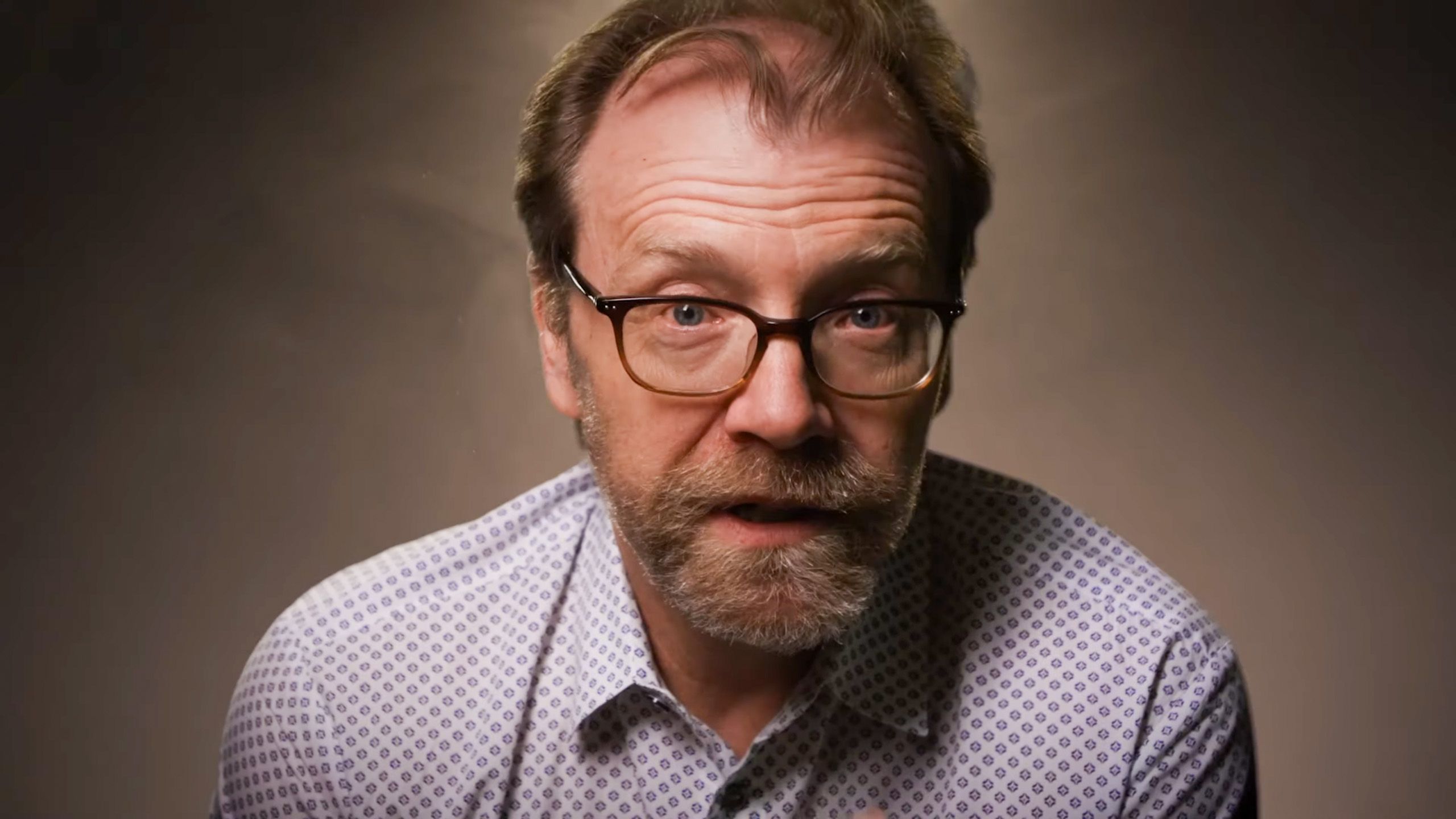 Photo of a man with glasses and a beard in a patterned shirt against a plain backdrop looking toward the camera.