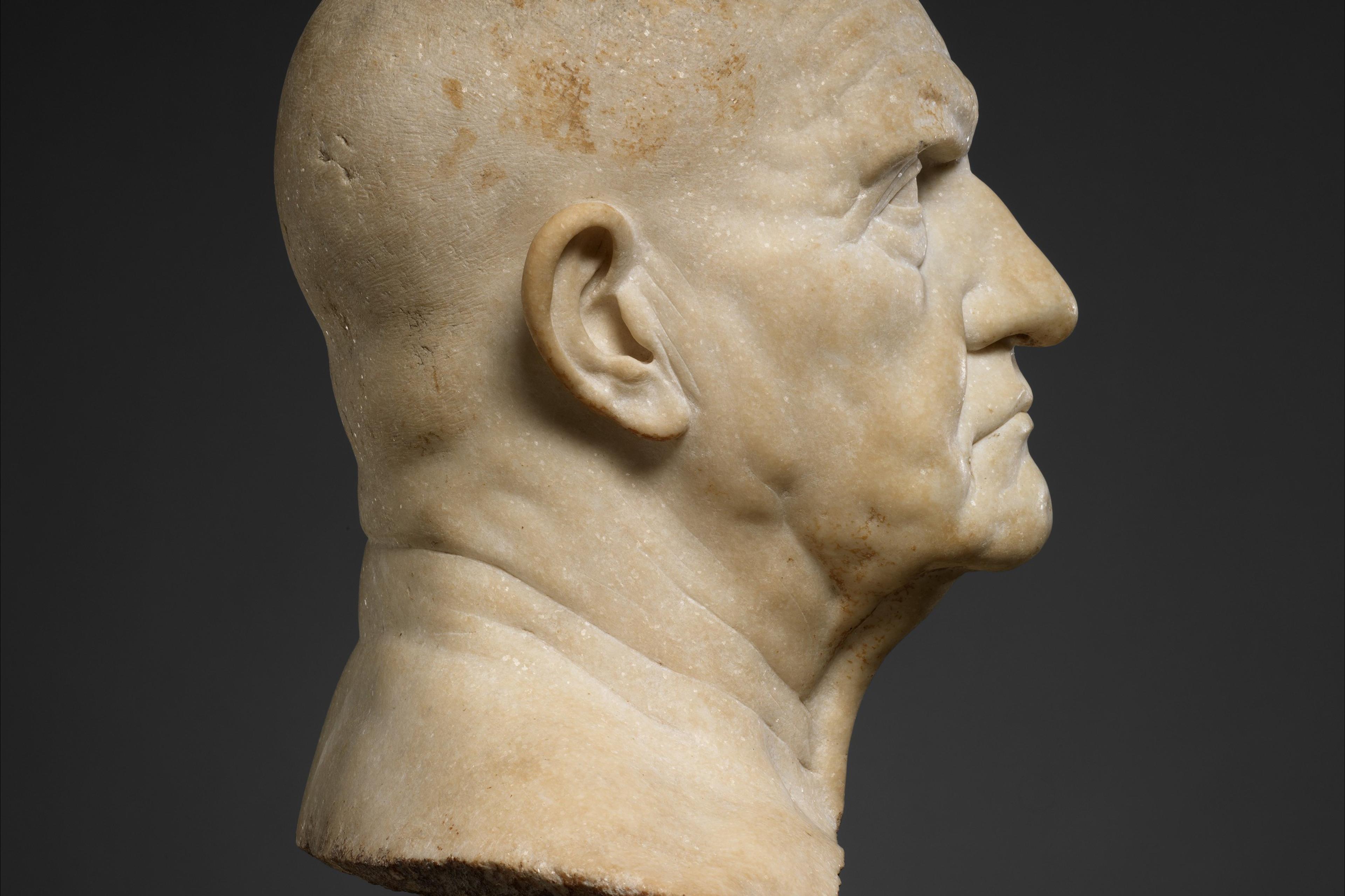 Side view of a marble bust of an elderly man with a stern expression on a dark background.