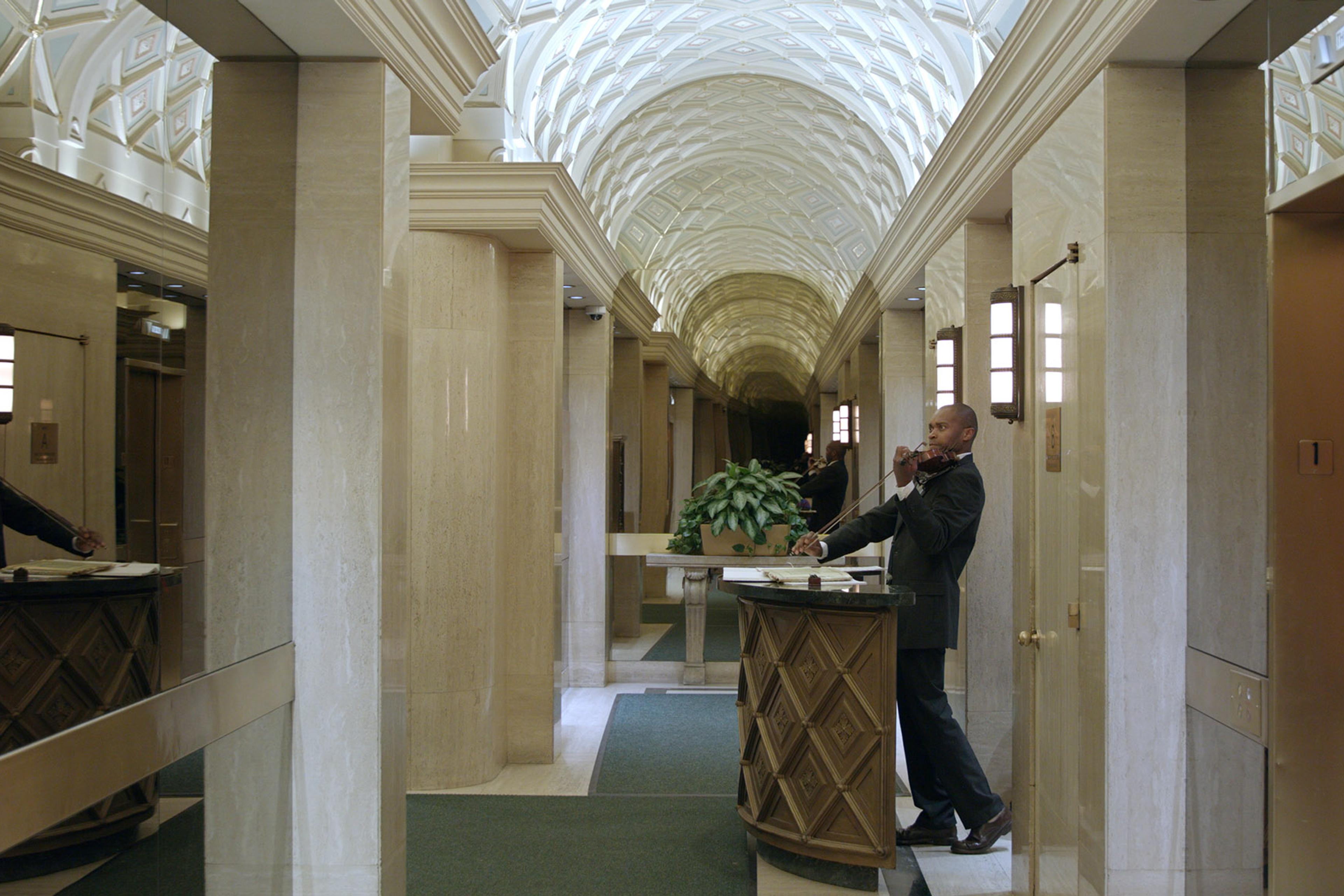 Man playing violin in stunning marble hallway with arched ceiling, elevators on both sides, plant in centre, reflections in glass.