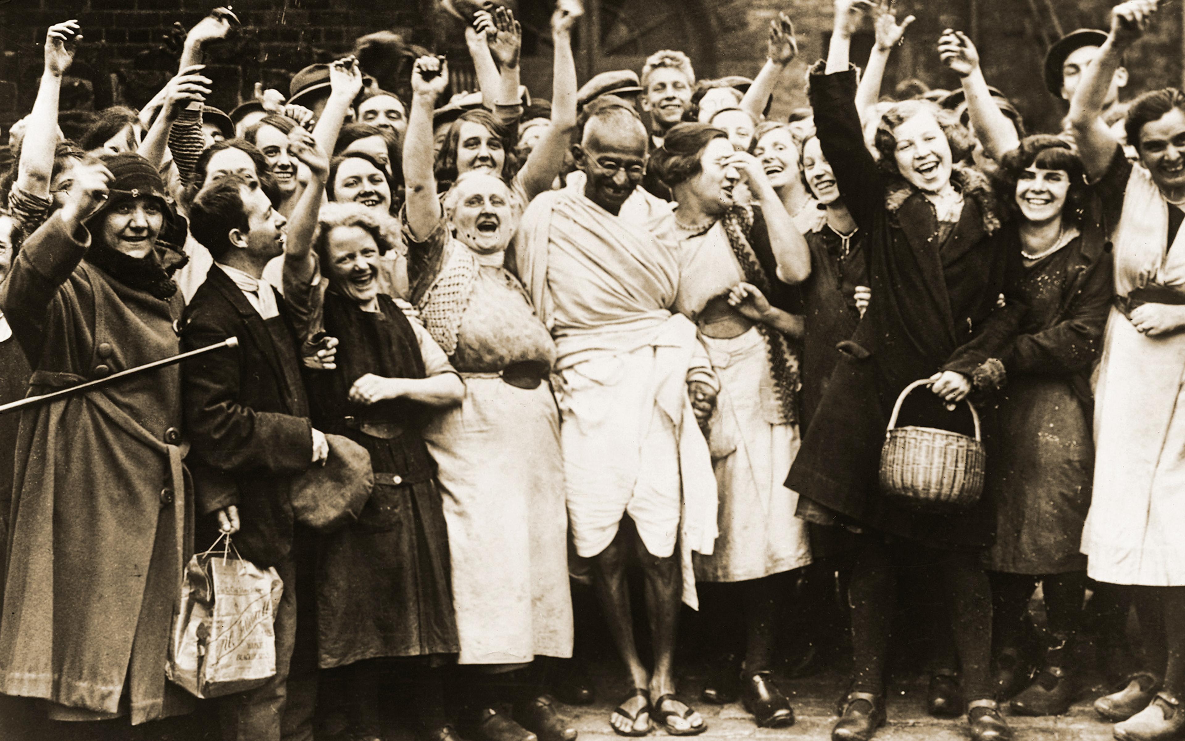 Black-and-white photo of a group of working people smiling and cheering with raised arms around a man in robes, sandals and glasses who is Gandhi.