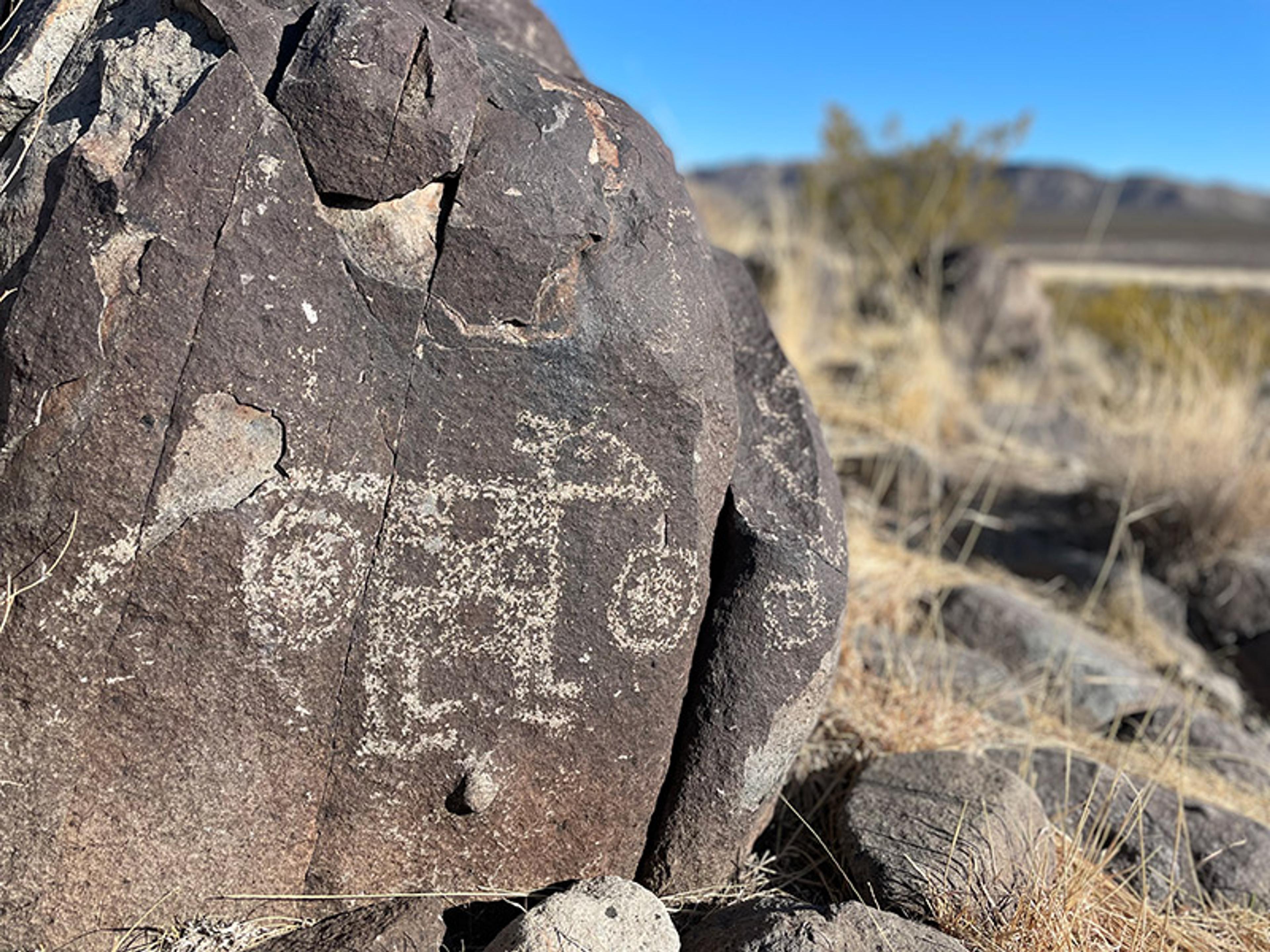 Ancient petroglyphs etched into a large rock in a desert landscape, with dry grass and distant mountains in the background.