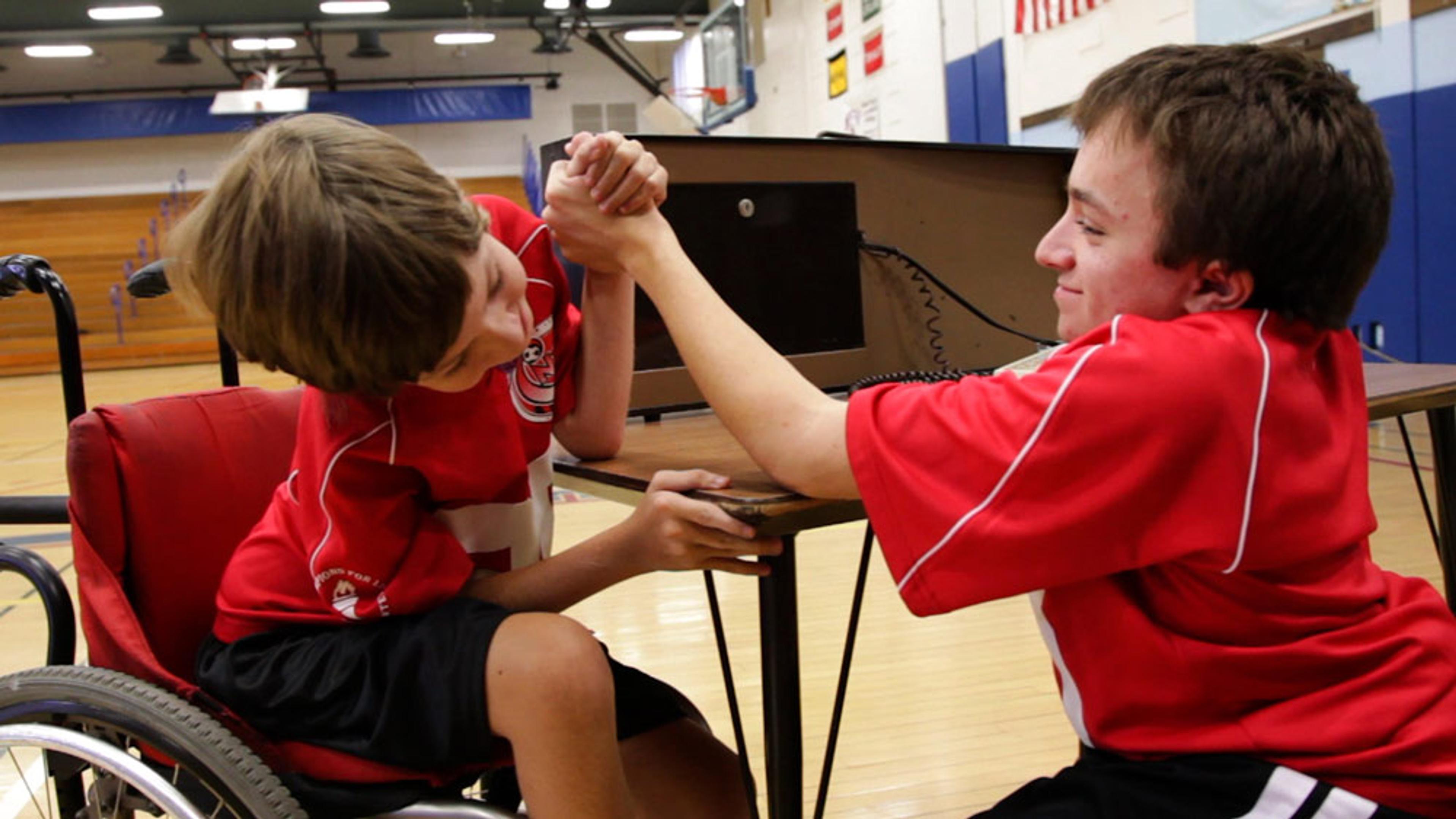 Two boys in red jerseys arm wrestling, one in a wheelchair, in a gym with wooden bleachers and flags in the background.