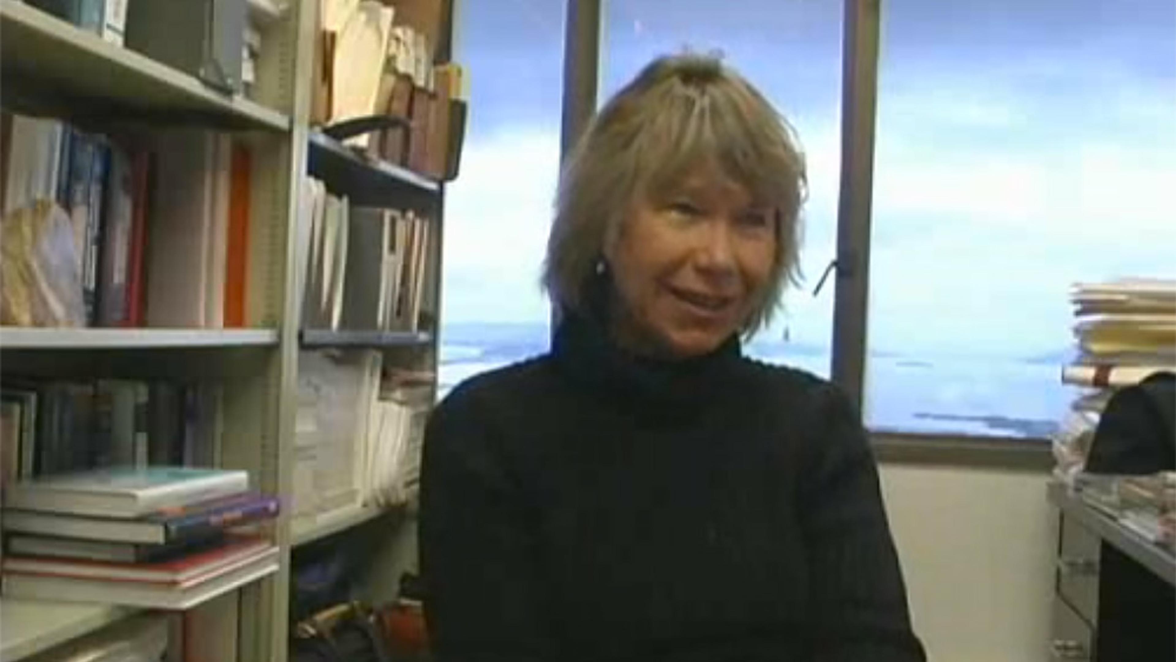 A woman with grey hair wearing a black jumper, sitting in an office with bookshelves and a window in the background.