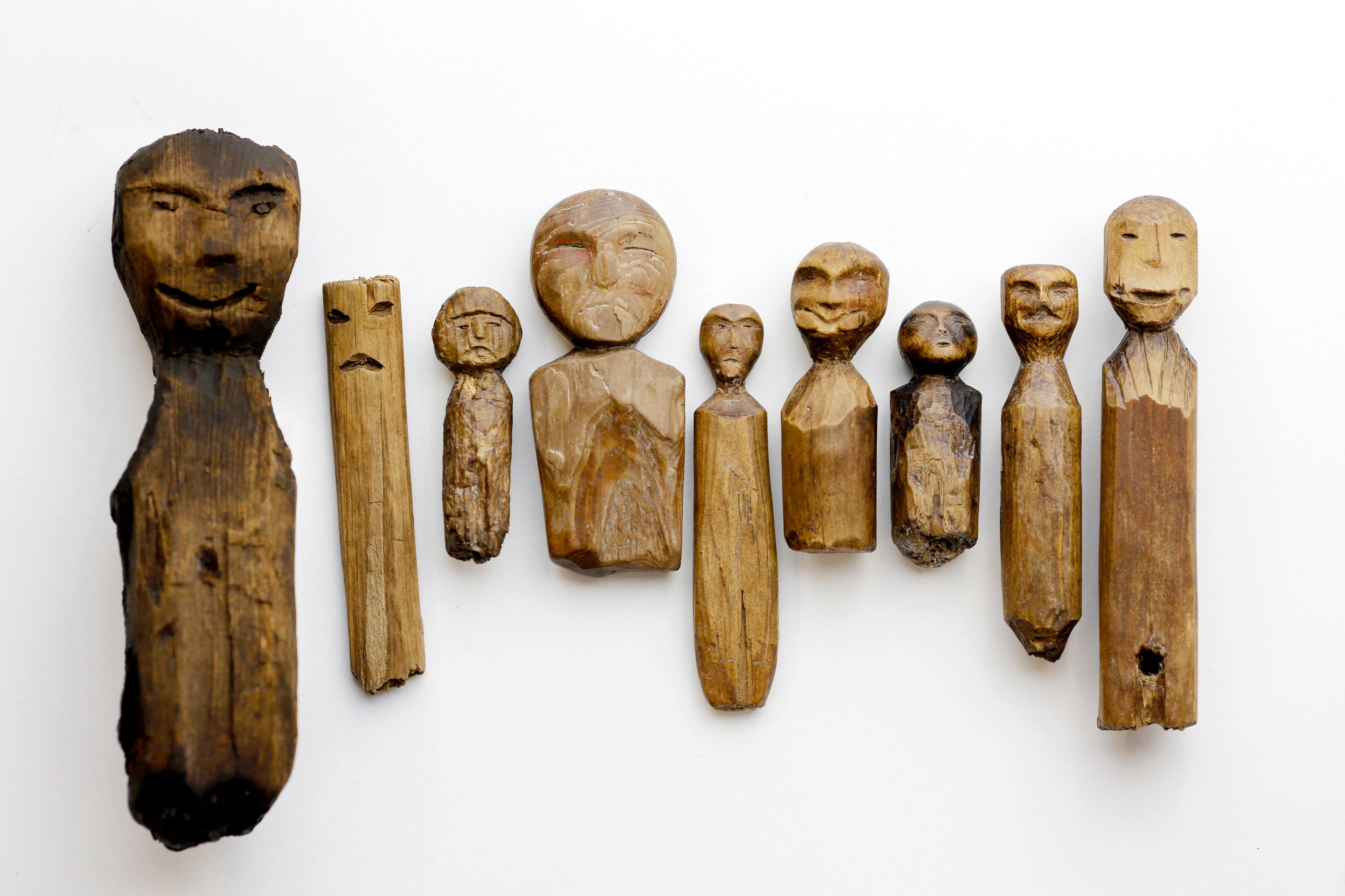 Photo of carved wooden figures on a white background each with simple facial features and varying shapes and expressions.