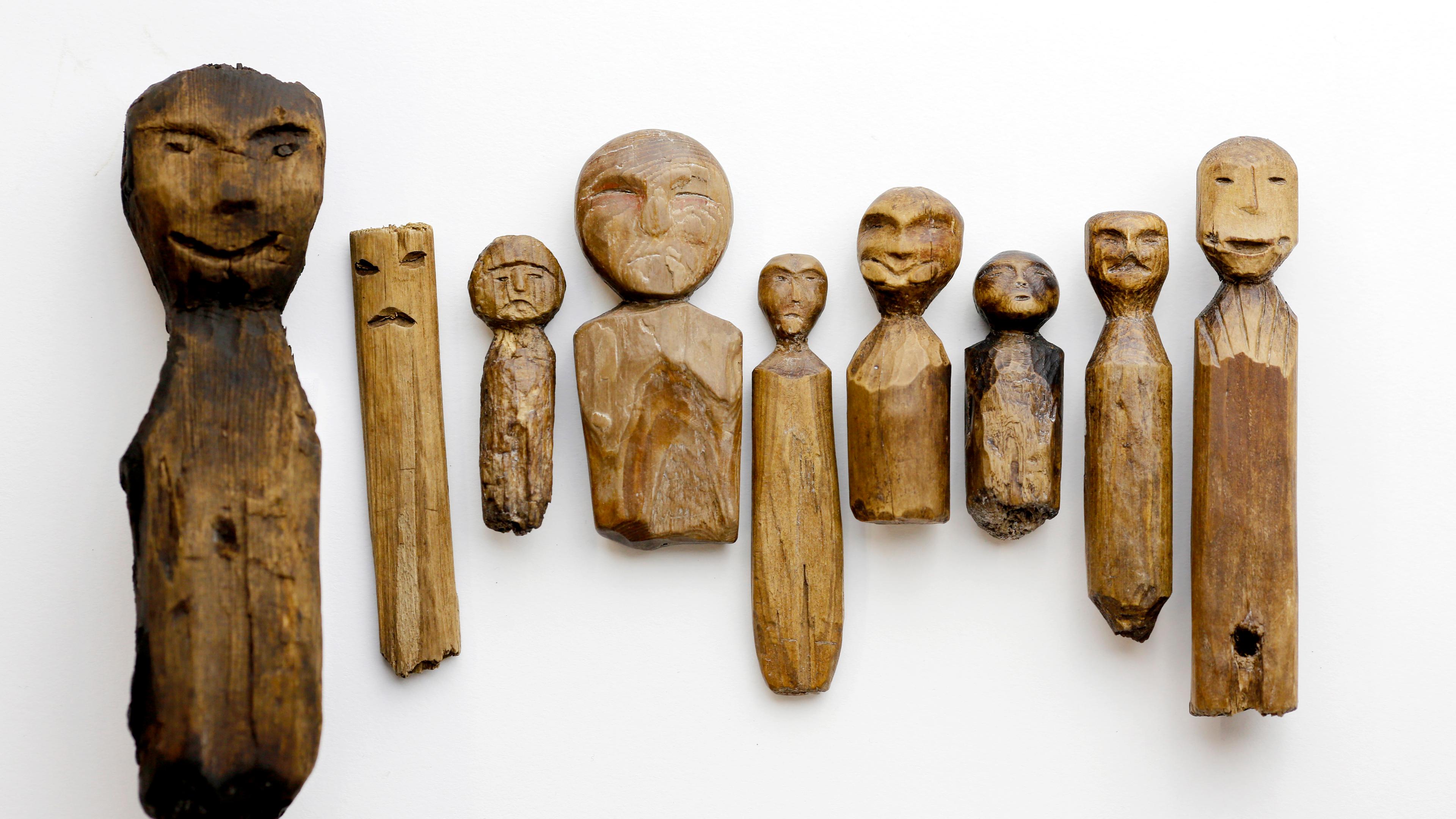 Photo of carved wooden figures on a white background each with simple facial features and varying shapes and expressions.