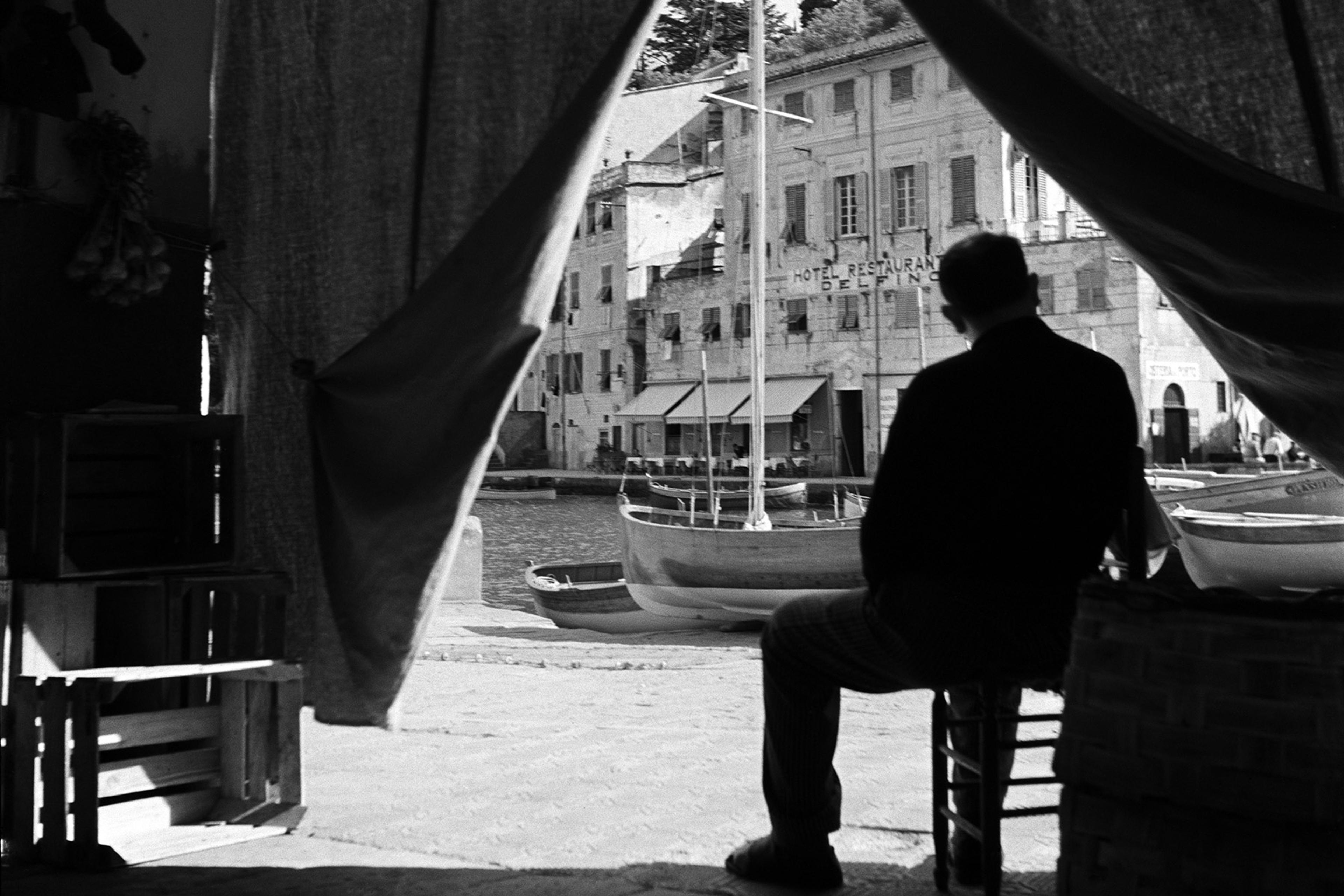 Black and white photo of a man silhouetted sitting in a doorway looking at boats and a hotel across a harbour.